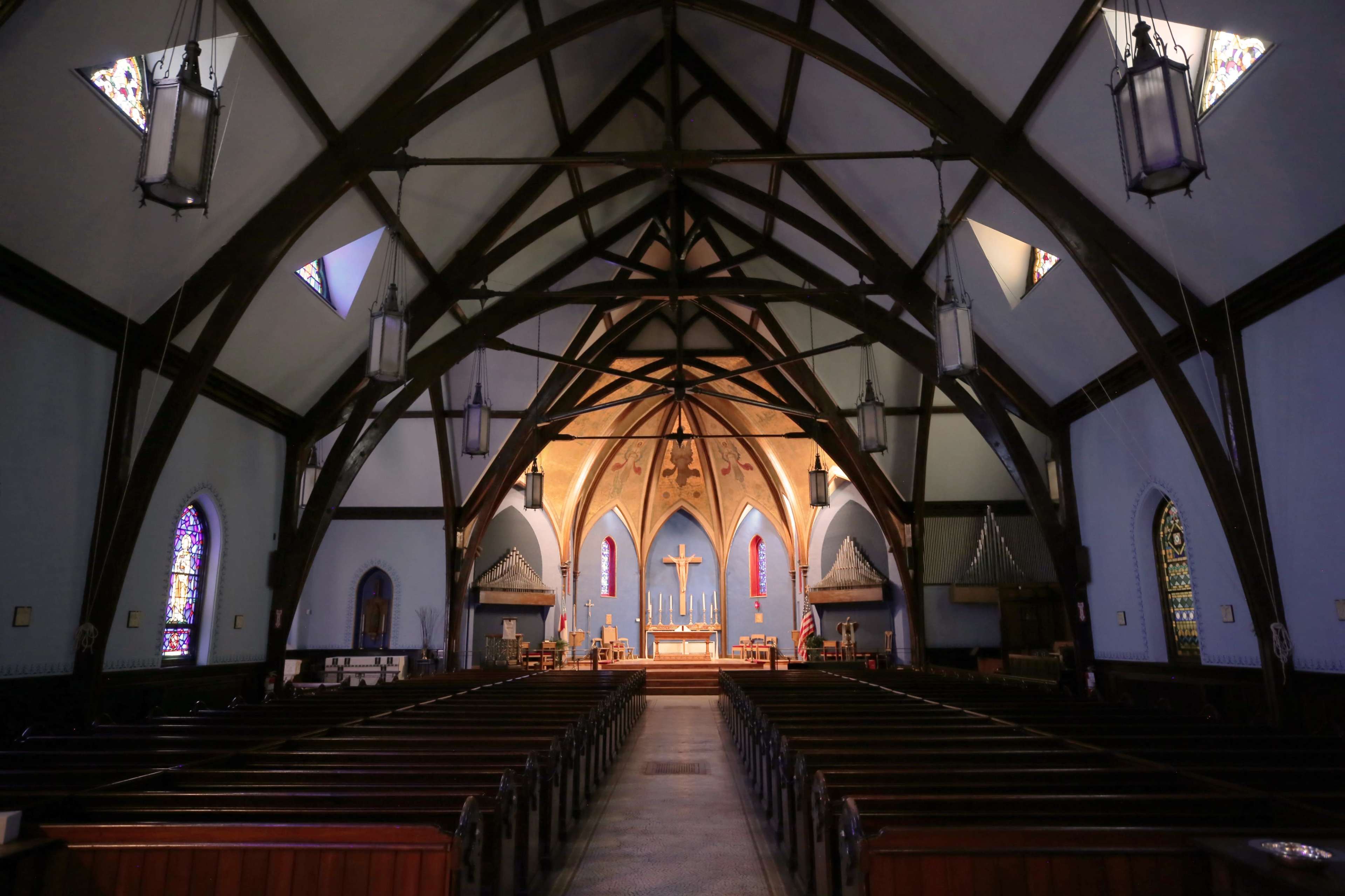 The interior of a church features a high, wooden vaulted ceiling, wooden pews arranged in rows, and stained glass windows along the walls.