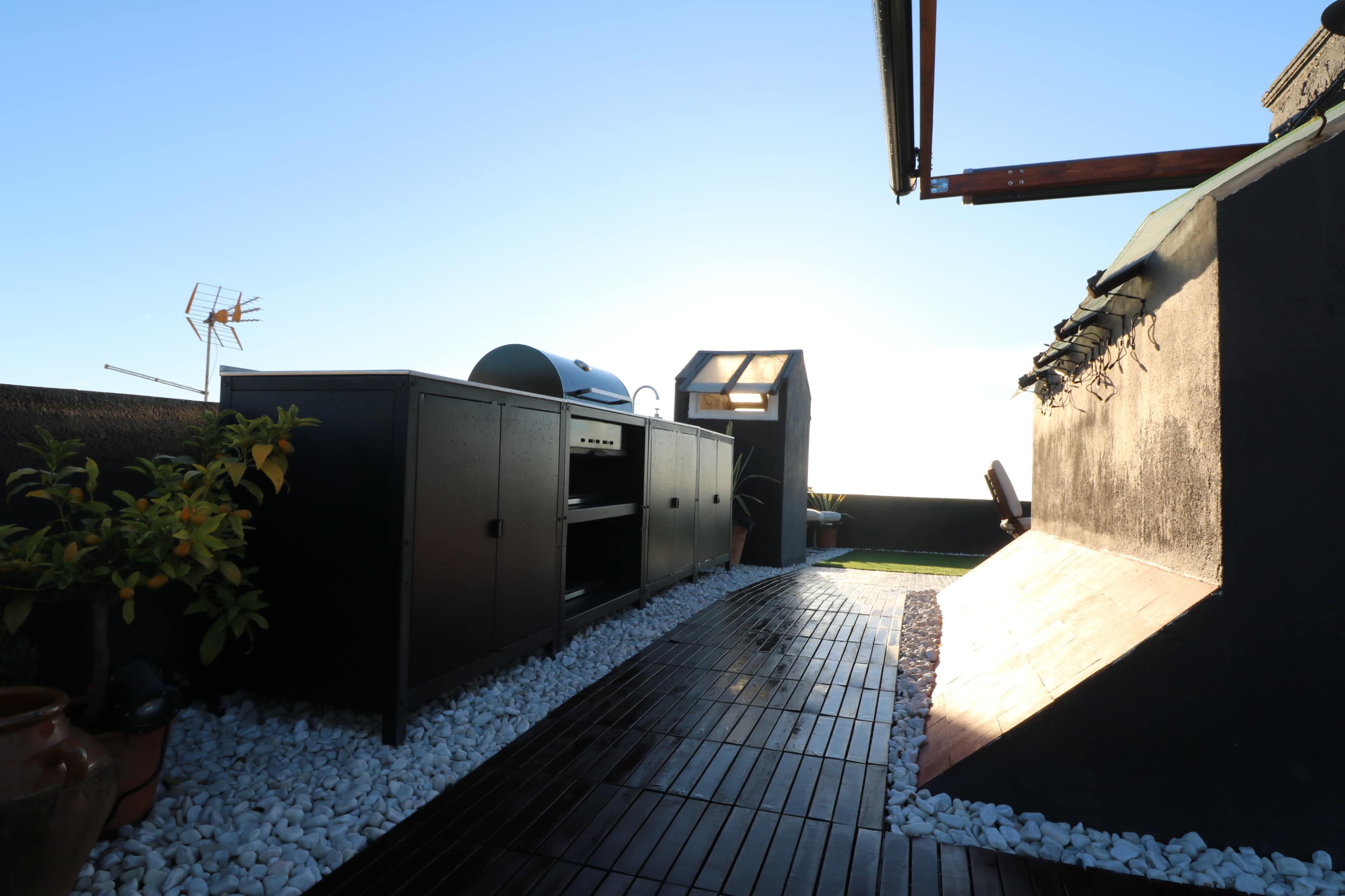 A rooftop space features a black outdoor kitchen unit with a grill, surrounded by white gravel and wooden decking.