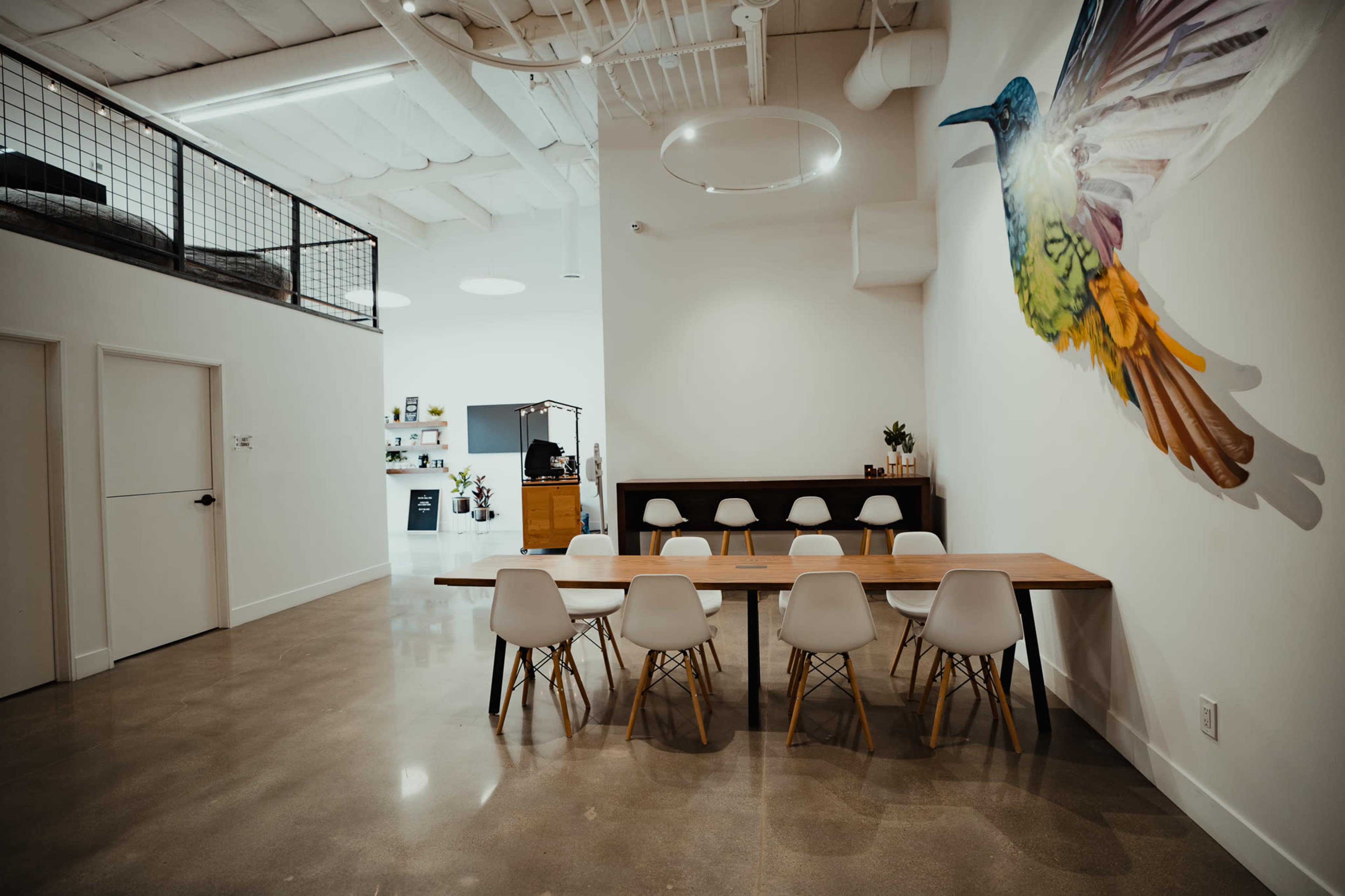 A modern, minimalistic dining area featuring a long wooden table surrounded by white chairs, with a colorful bird mural on the wall and a clean, open space.