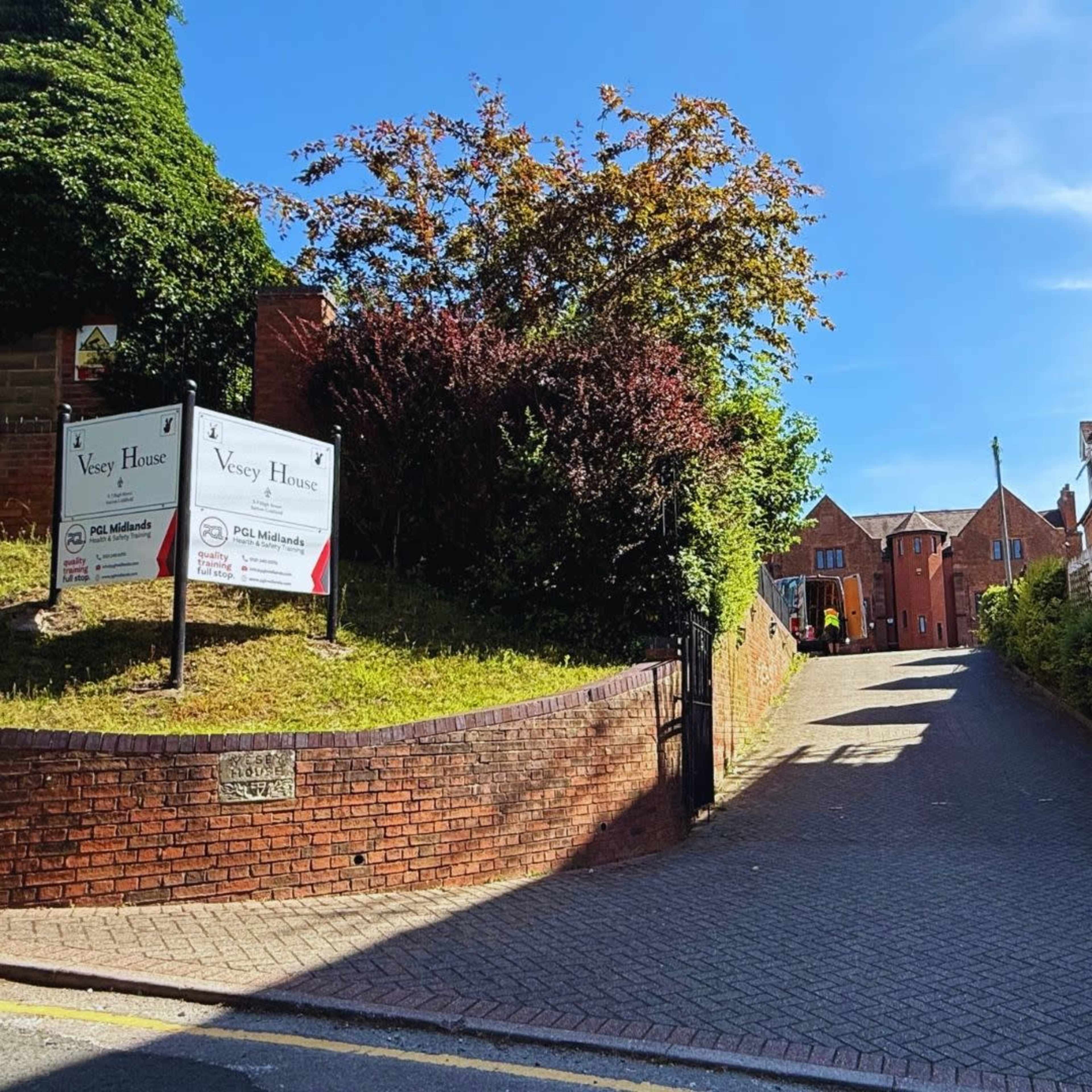 The image shows a brick pathway leading up a hill, flanked by greenery and signs for "Vesey House" on the left.