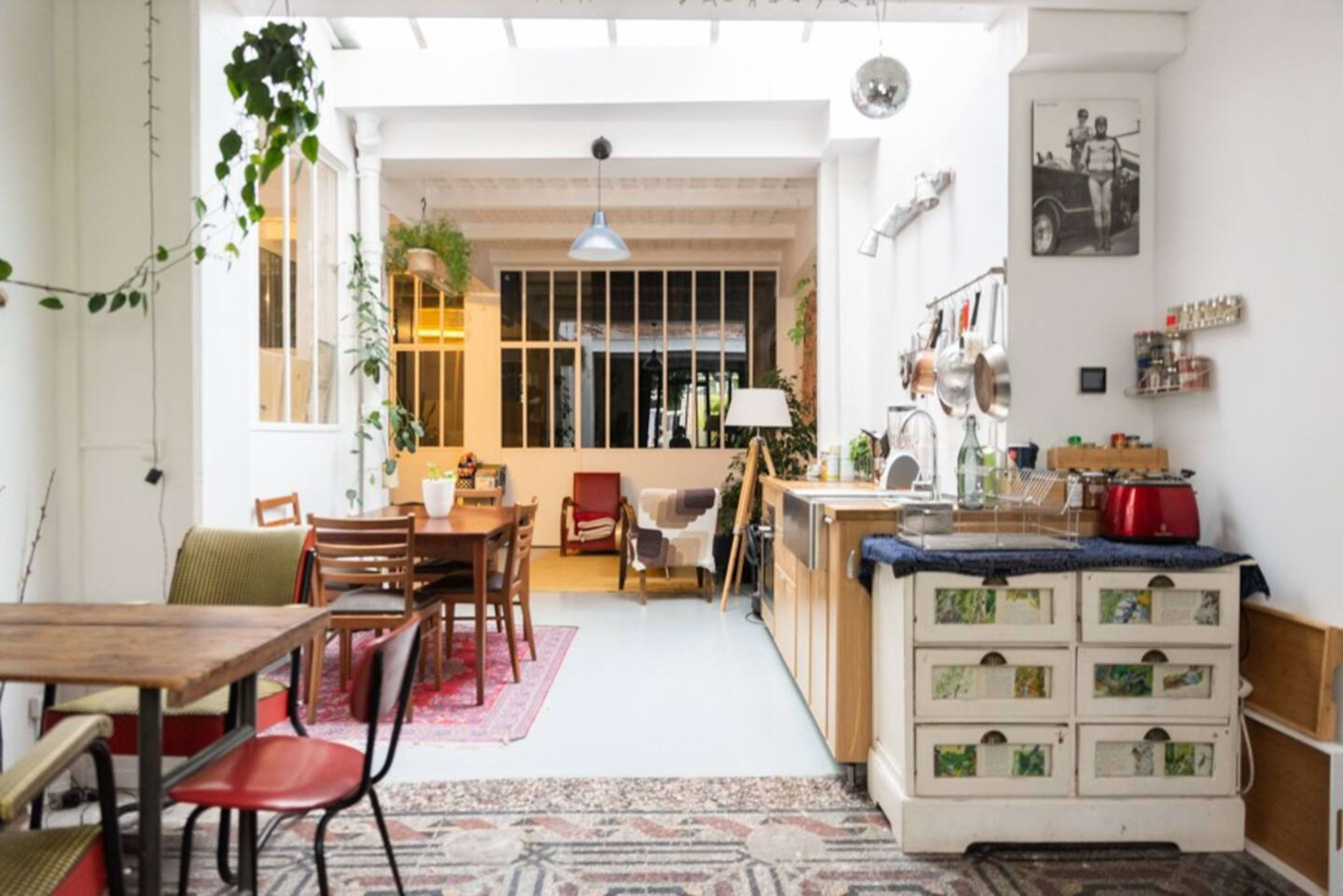 The image depicts a spacious kitchen and dining area with wooden tables, chairs, and various plants around large windows.