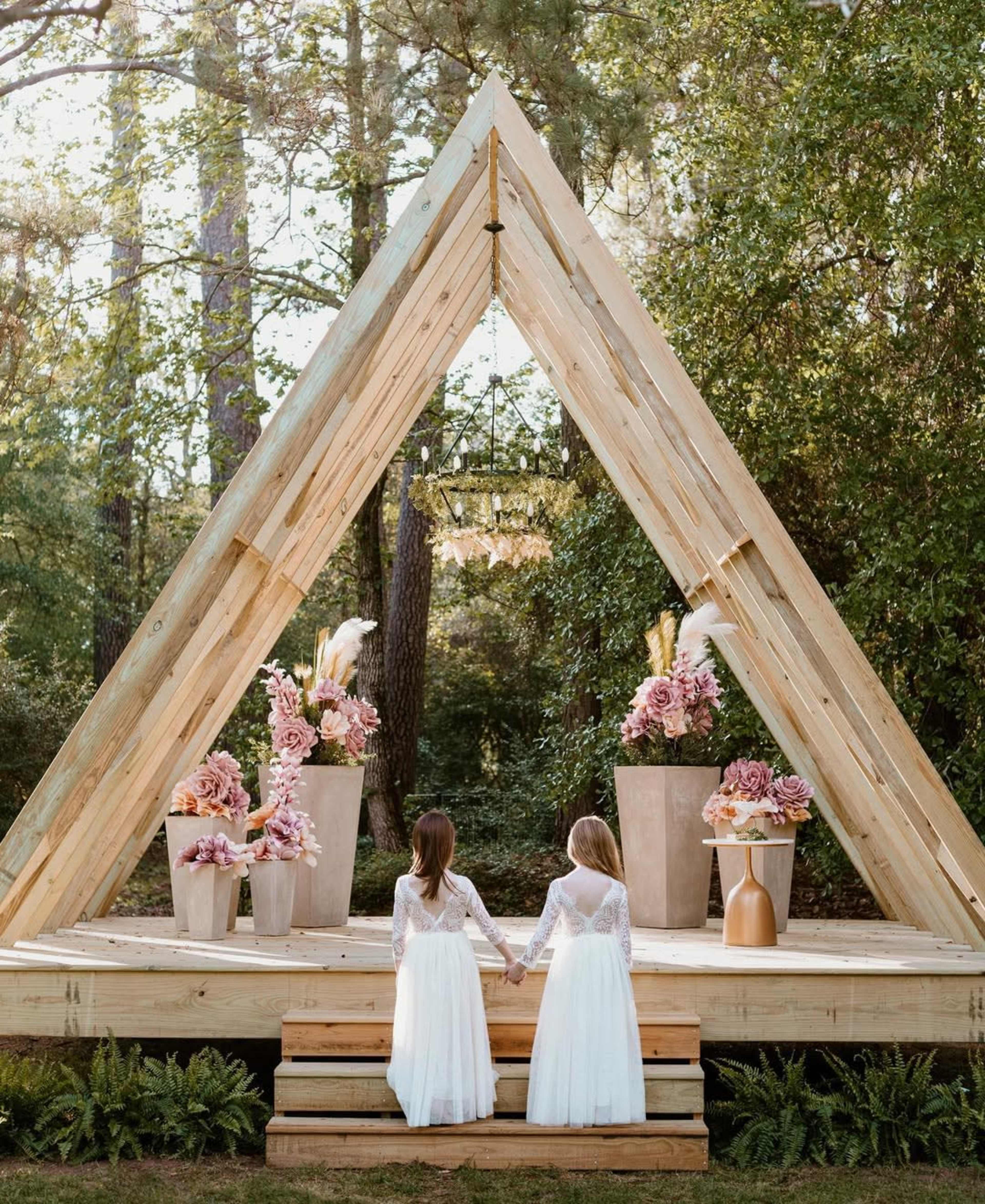 Two girls in white dresses stand hand-in-hand on wooden steps in front of a triangular wooden structure adorned with floral arrangements.