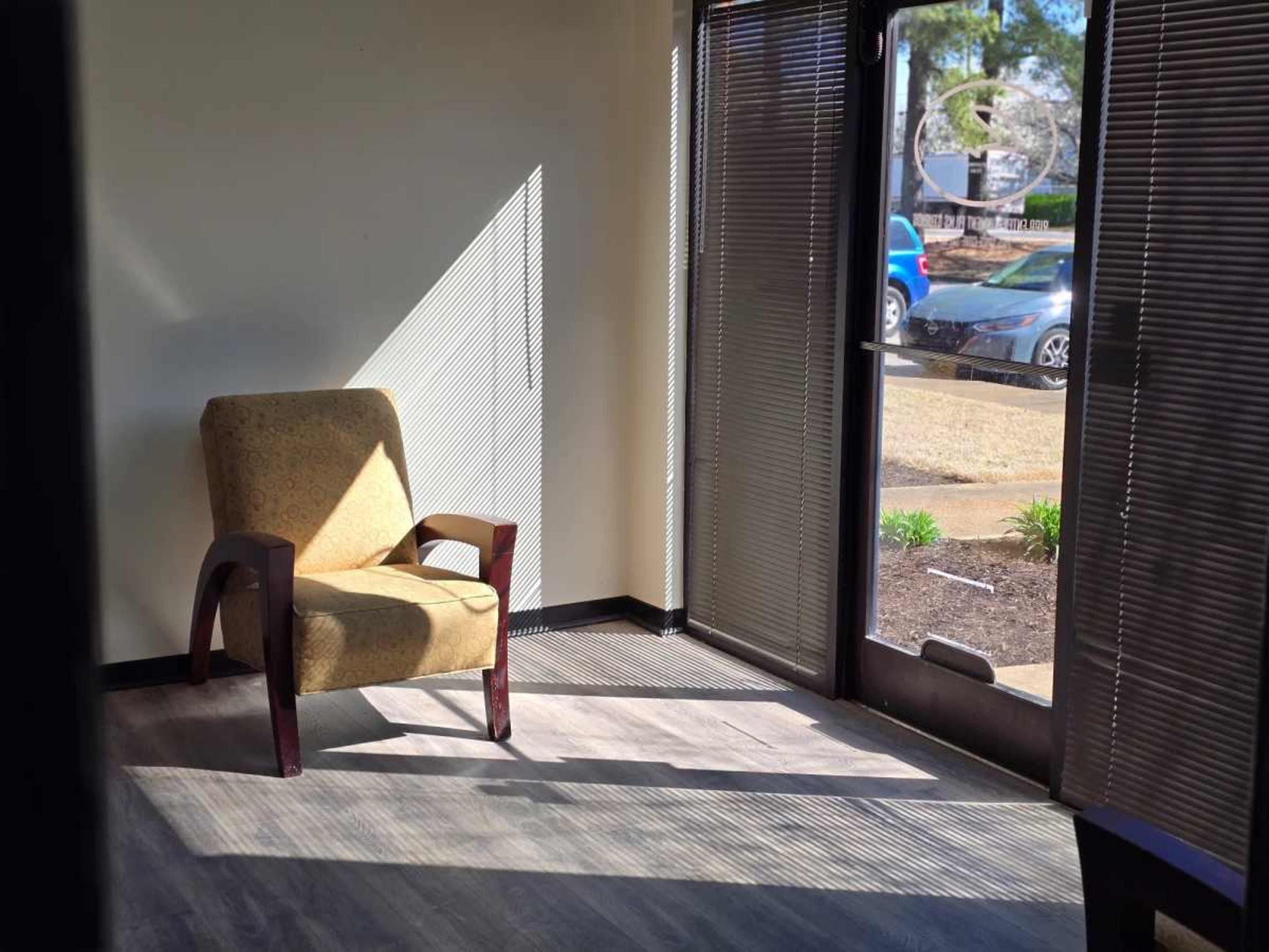 A single upholstered chair is positioned in a sunlit corner of an office with large windows and blinds cast shadows on the floor.
