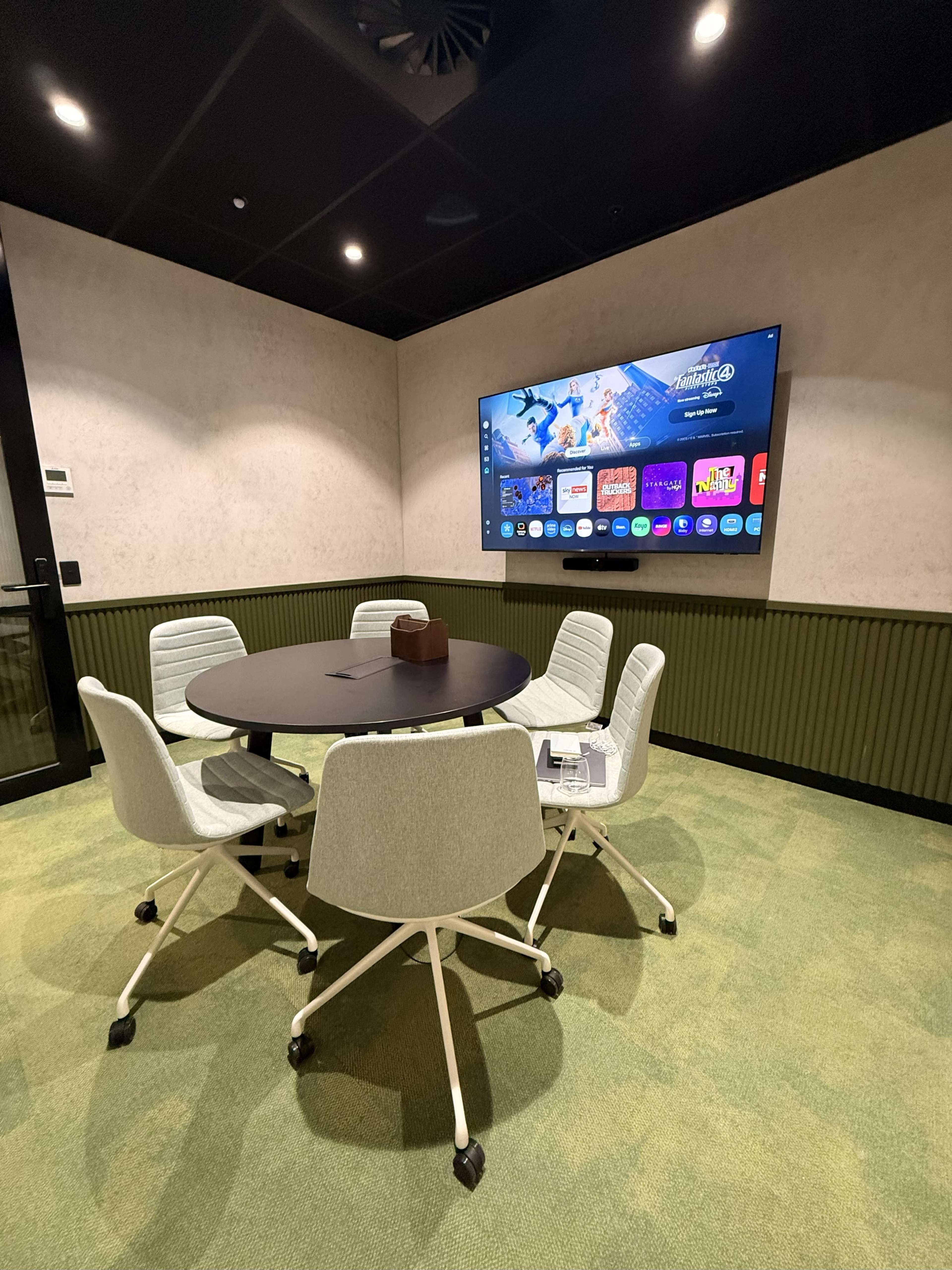 A conference room features a round table surrounded by white chairs, with a large screen displaying apps on a wall.