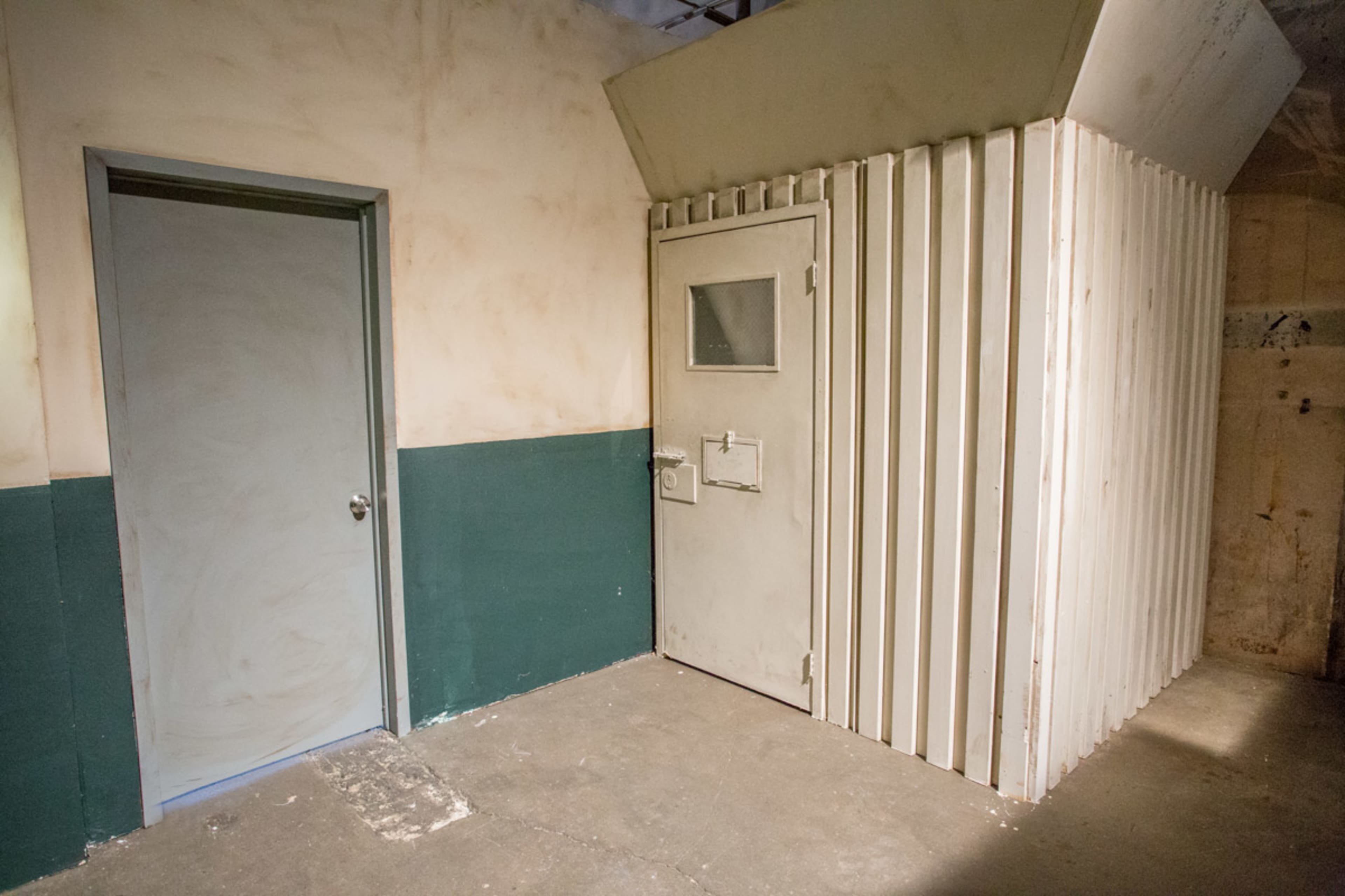 A small room with a plain door on the left and a secure, metal door with a window on the right, both against a textured wall painted in muted colors.