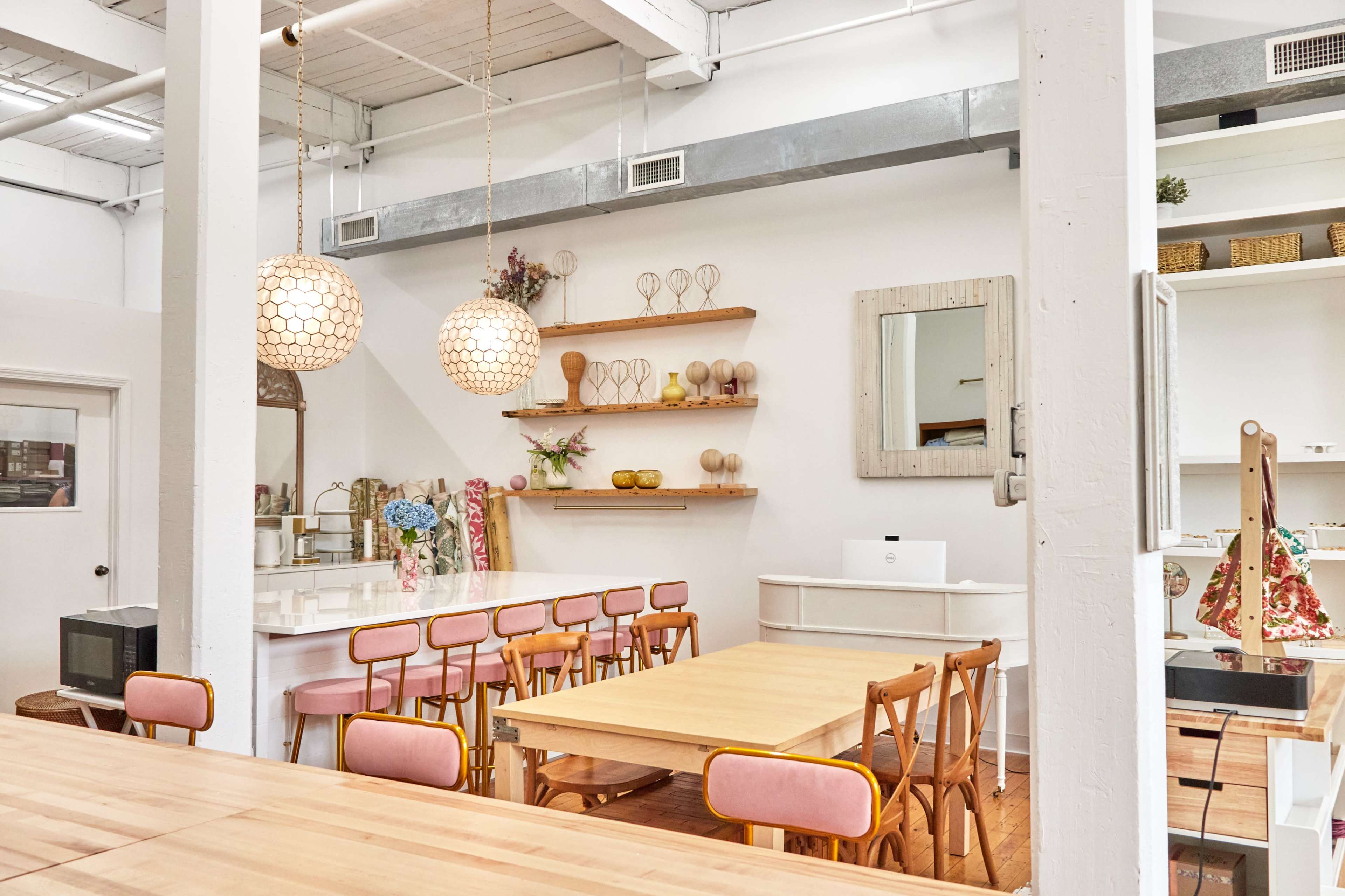 A bright, modern interior of a café or shop featuring wooden tables, pink chairs, and decorative shelves with various items displayed.