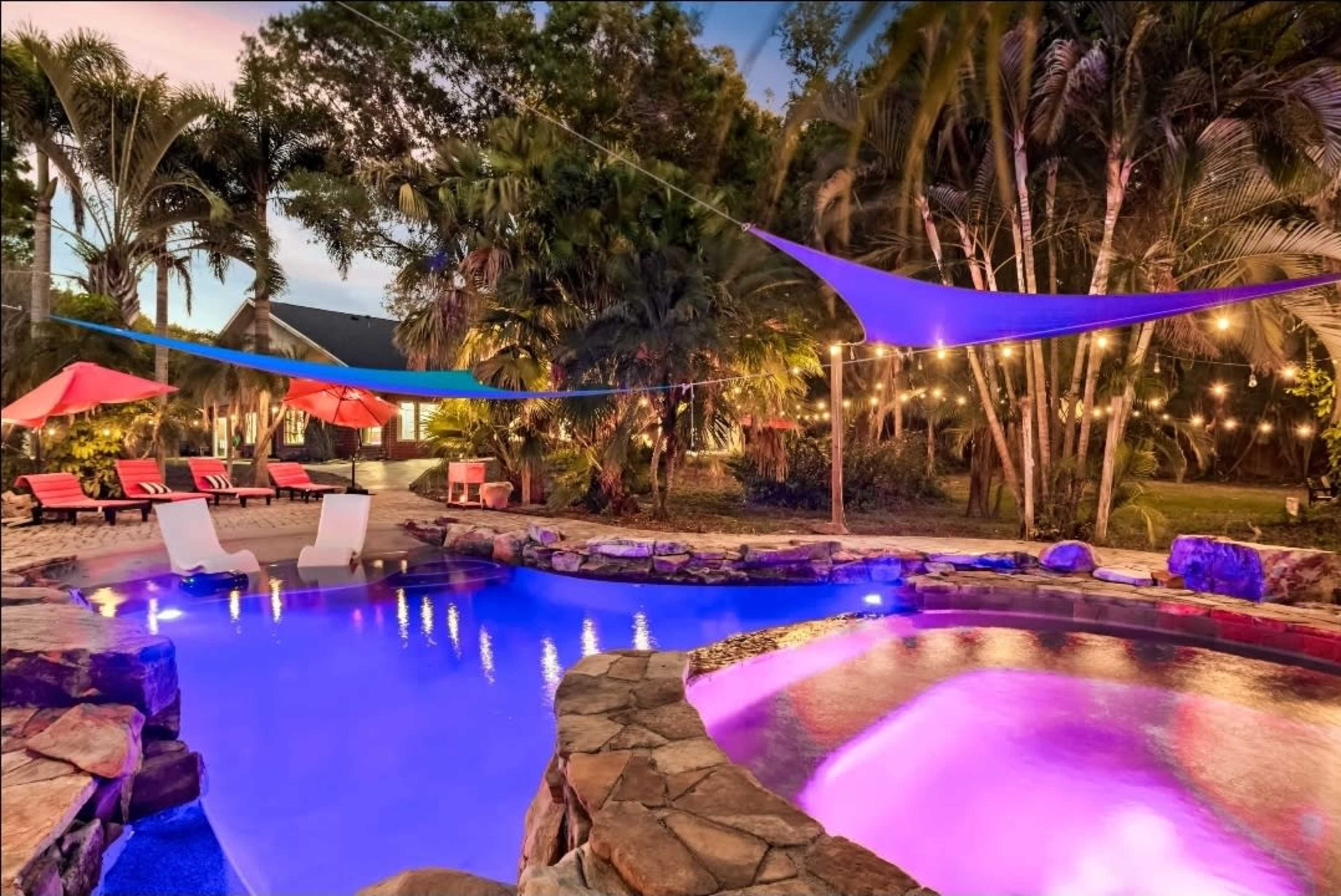 A vibrant outdoor pool area features colorful lounge chairs, illuminated water, and decorative lighting amidst tropical foliage.