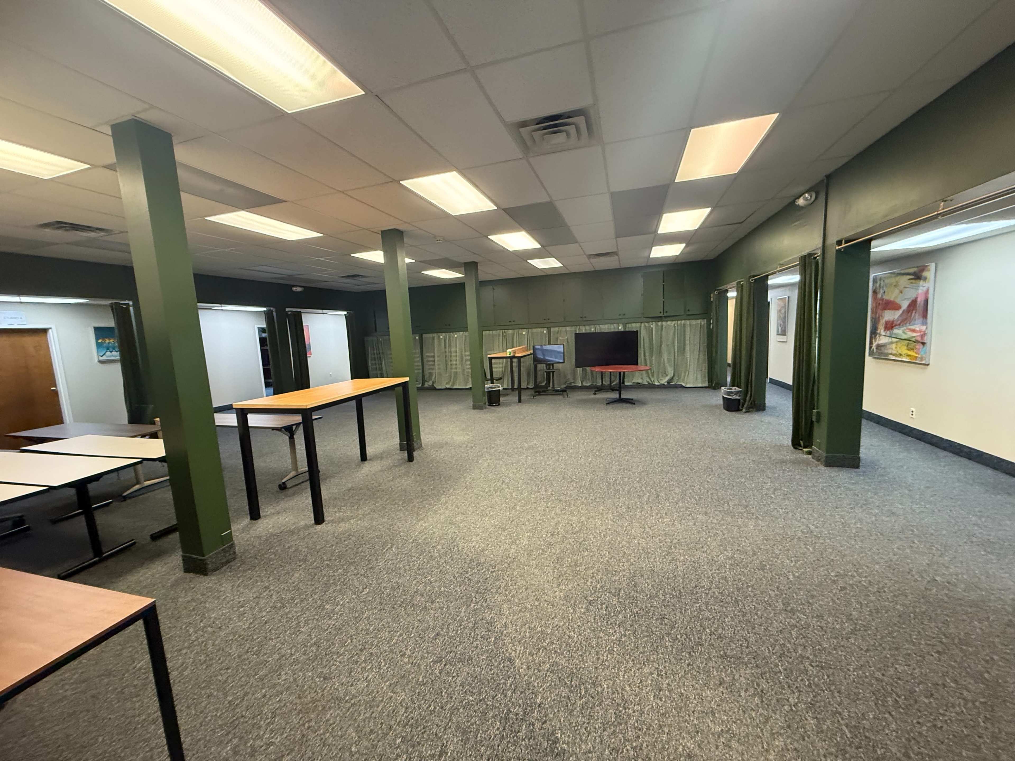 The image shows a spacious, empty room with green pillars, several tables, and a presentation screen in the center.