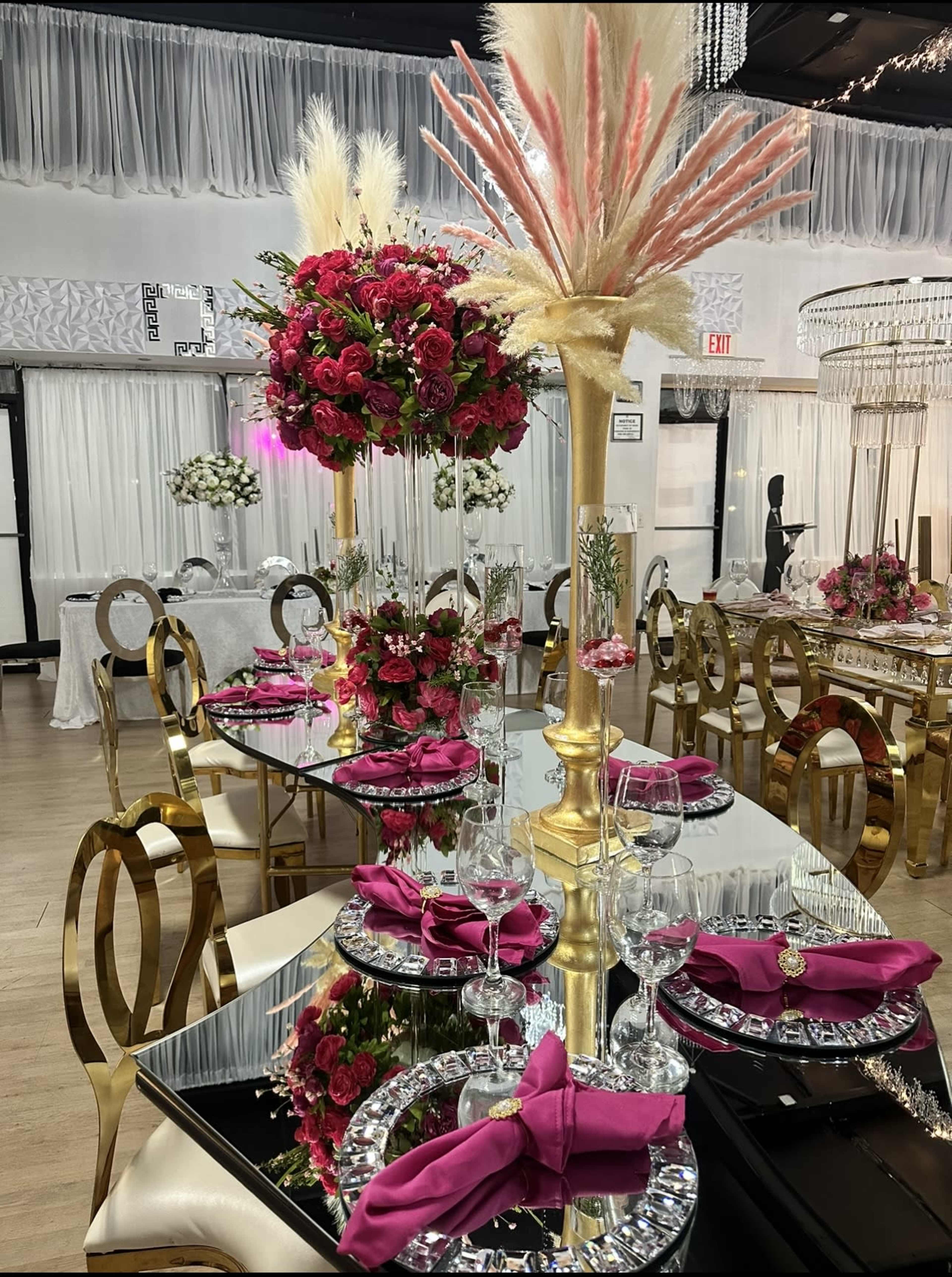 A beautifully arranged dinner setting with tall floral centerpieces, gold-accented tableware, and elegantly folded pink napkins on mirrored tables.