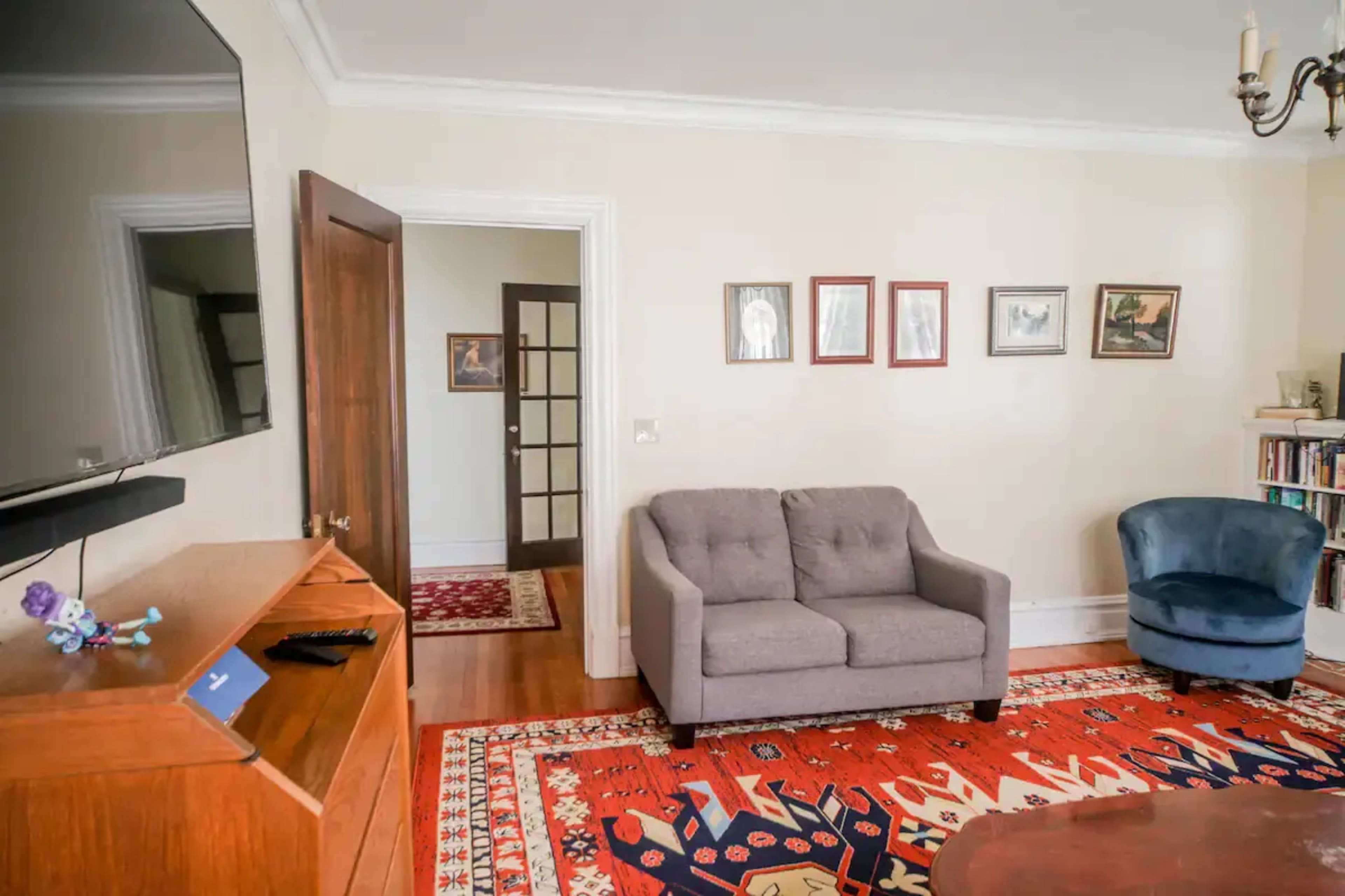The room features a gray sofa and a blue armchair, with framed artwork on the wall opposite a wooden furniture piece and a doorway leading to another area.