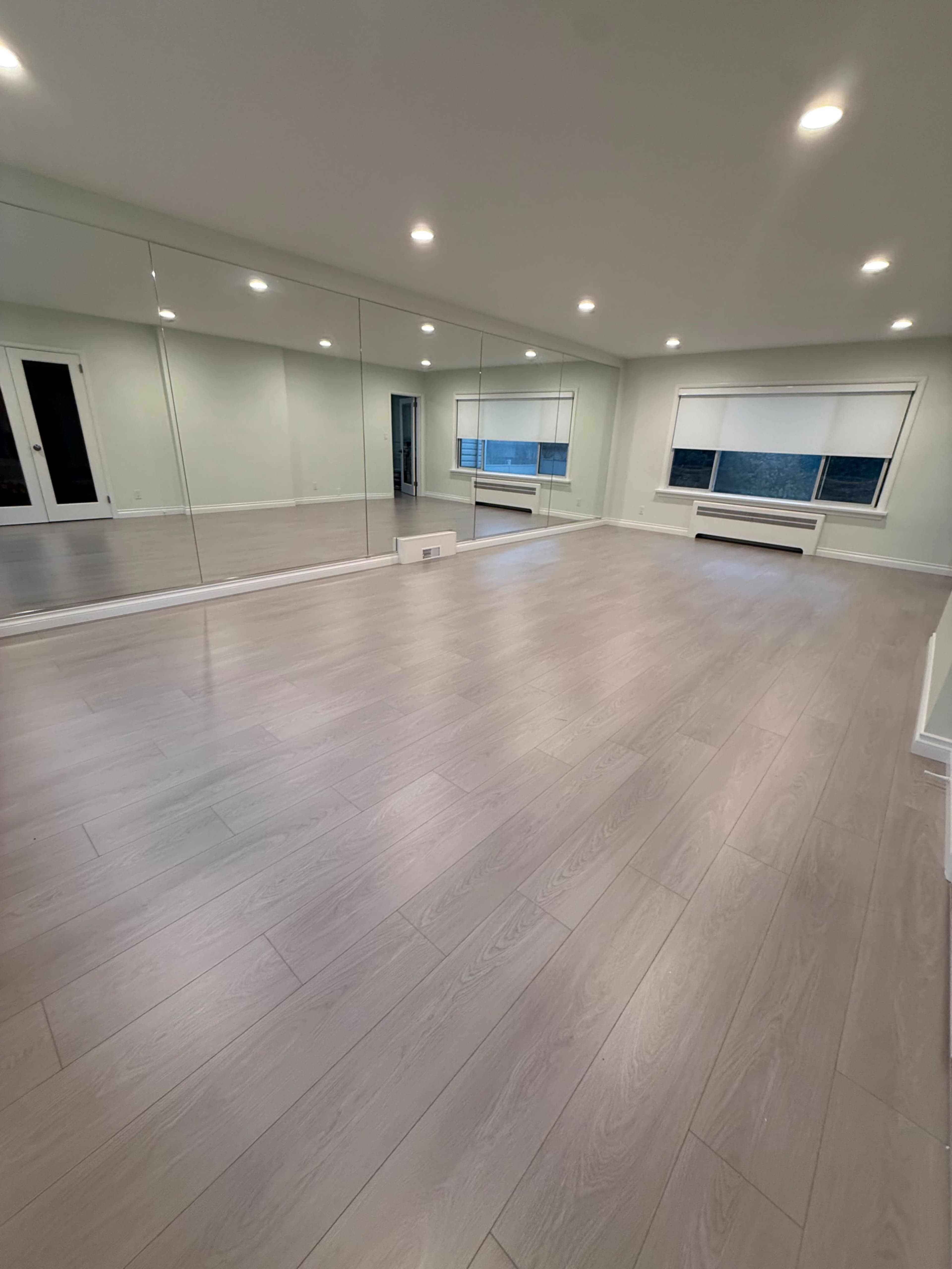The image shows a spacious, empty room with large mirrored walls and hardwood flooring, illuminated by overhead lights.