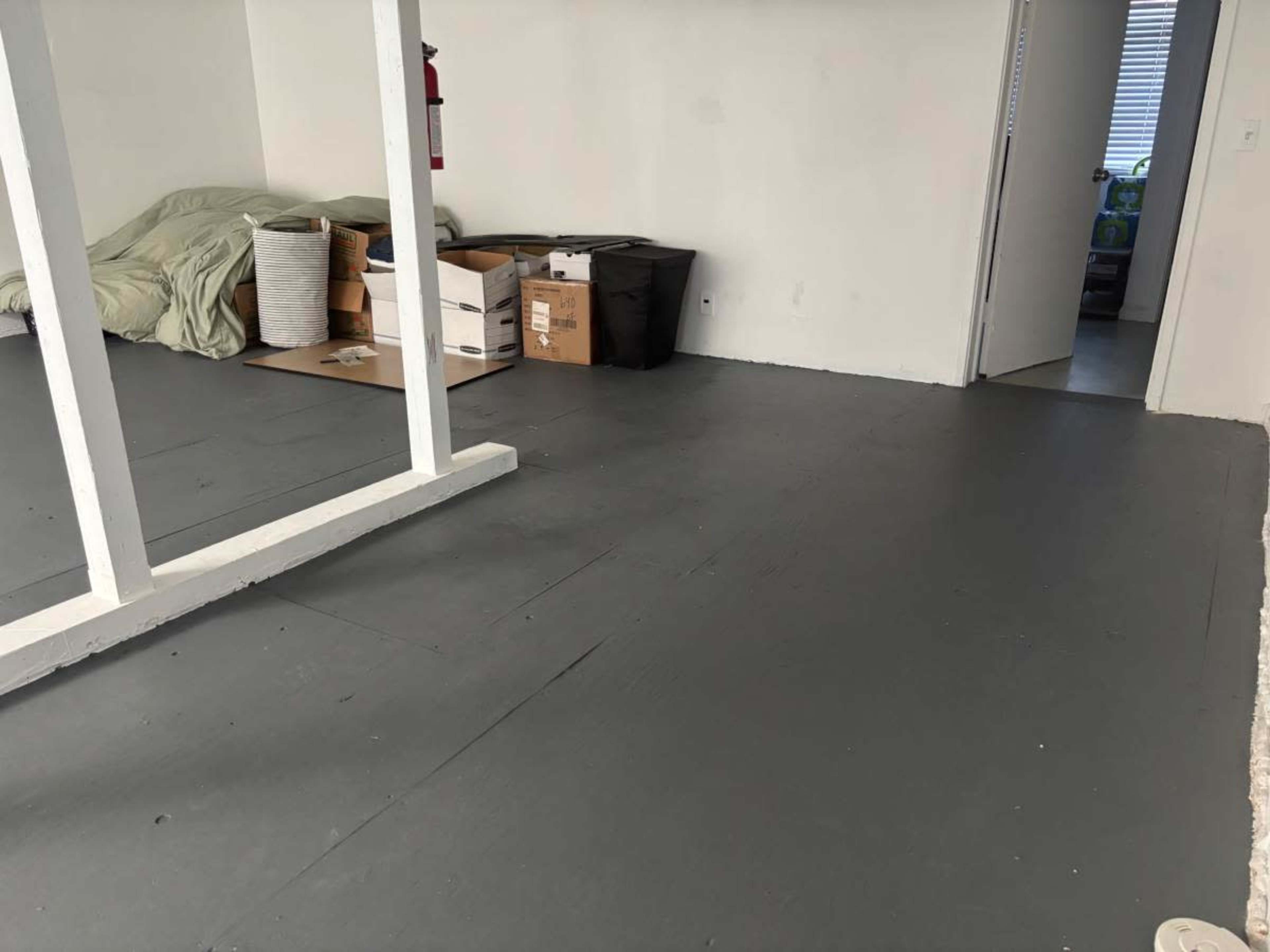The image shows a minimal interior space with a gray floor, several cardboard boxes, a trash bin, and an unmade bed in the corner.