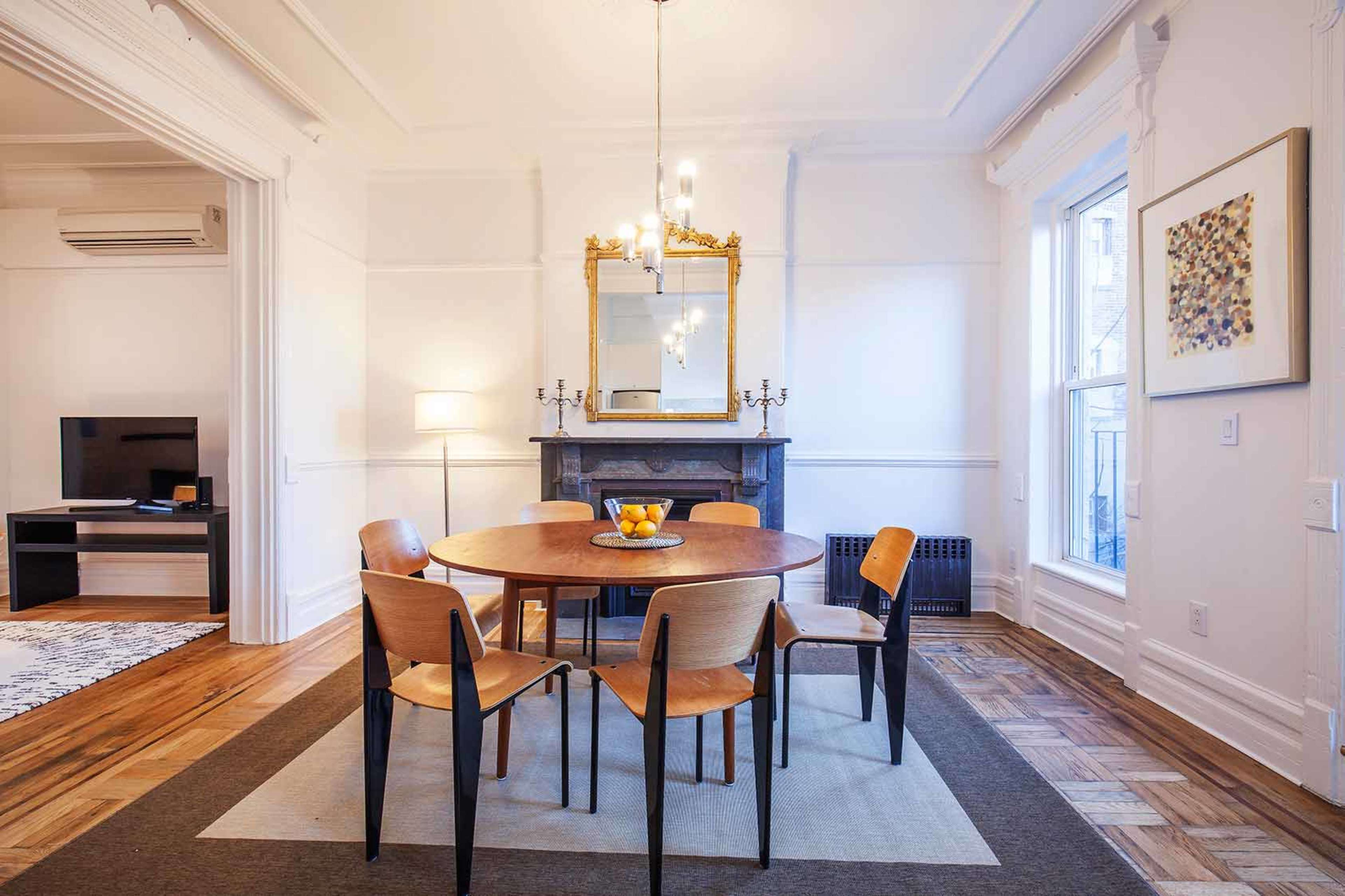 The image shows a dining area with a round wooden table surrounded by four chairs, a decorative mirror, and a fireplace in a well-lit room.