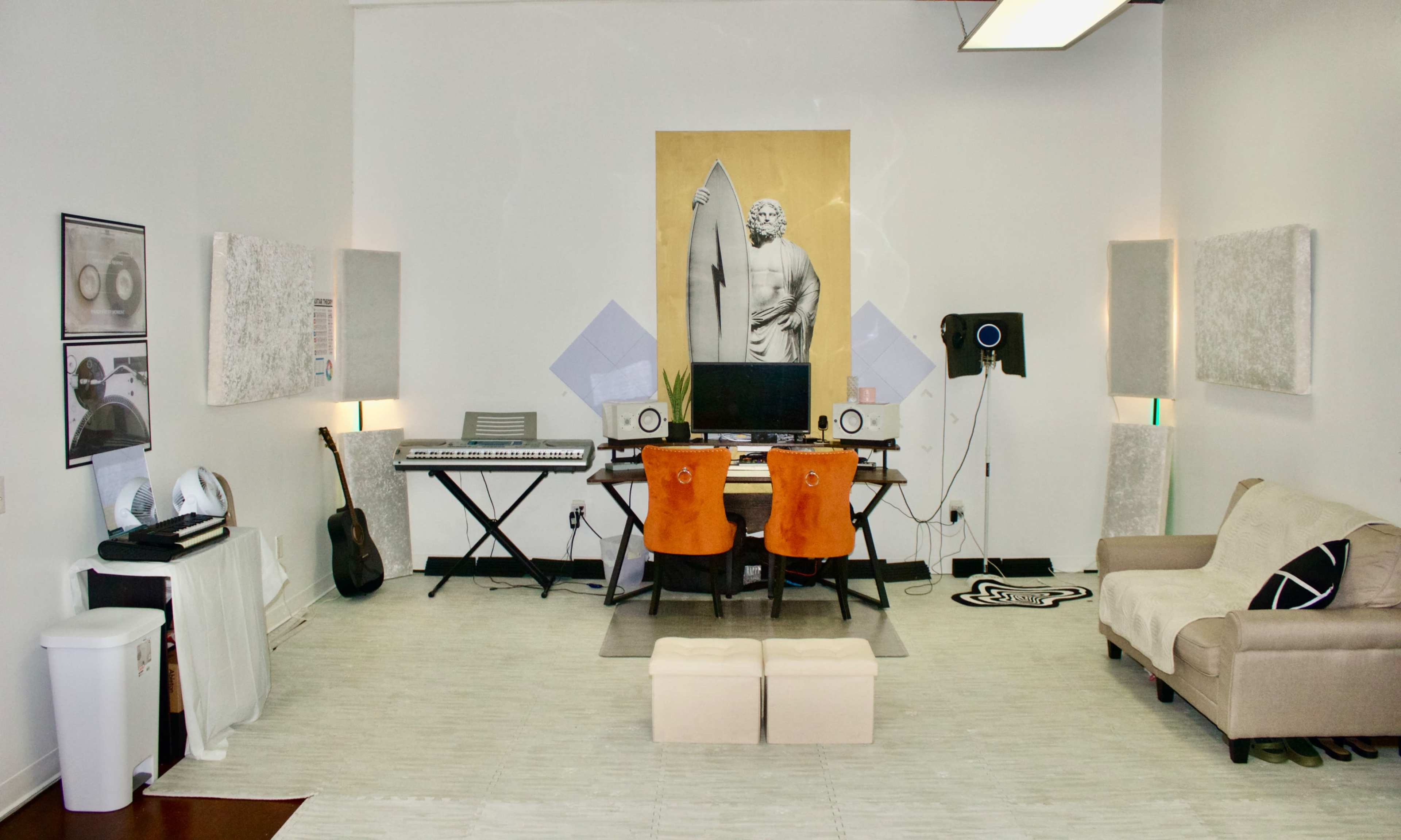 The image shows a stylish studio space featuring a desk with two orange chairs, a keyboard, a computer monitor, and a guitar, along with soundproofing panels on the walls and a beige couch in the corner.