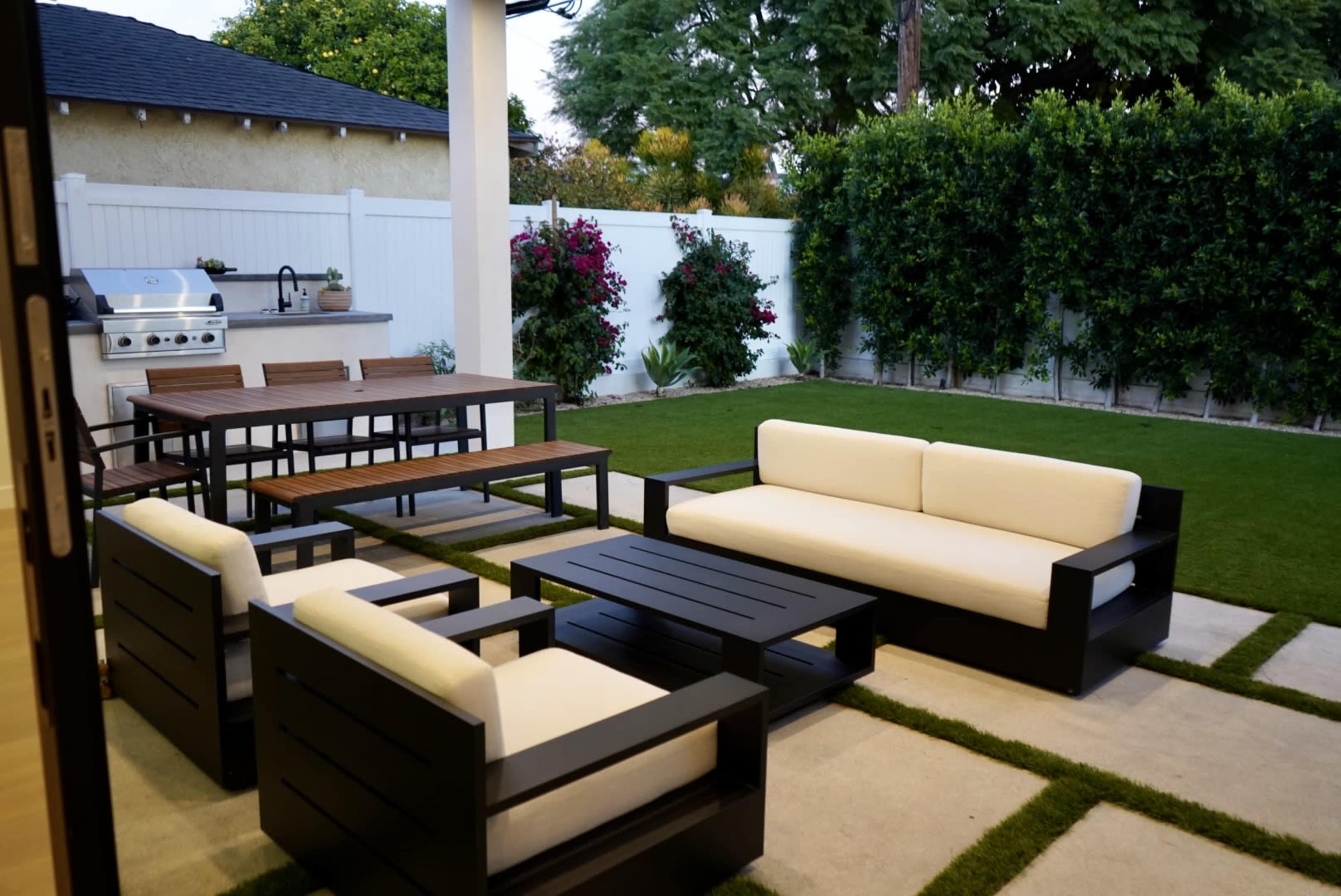 The image shows an outdoor patio with a seating arrangement of black wooden furniture, a dining table, and a barbecue grill against a backdrop of hedges and flower-filled bushes.