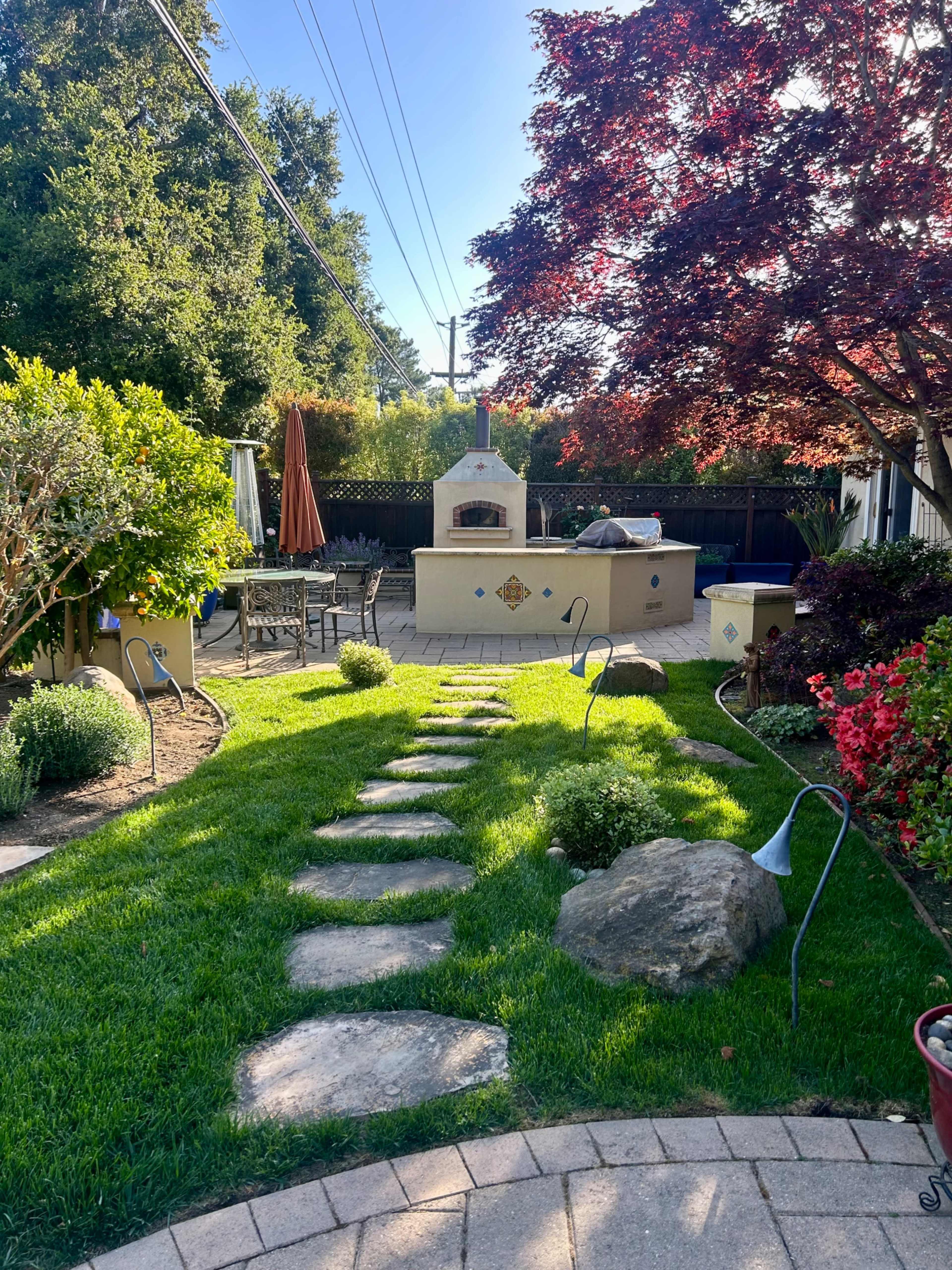 A landscaped backyard with stone pathways leading to an outdoor pizza oven, surrounded by greenery and patio furniture.