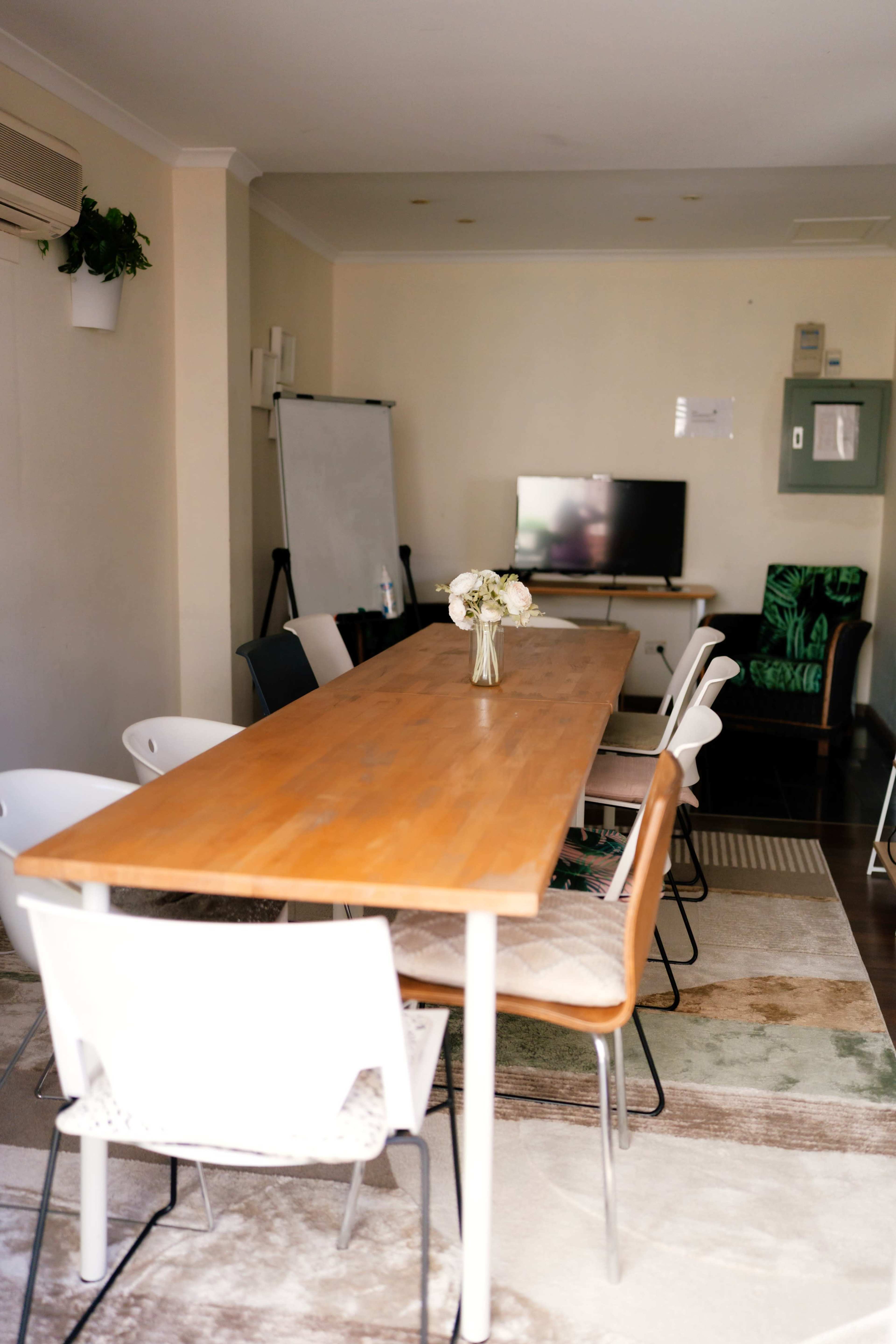 The image shows a conference room with a wooden table surrounded by white chairs, a television screen, a whiteboard, and a floral centerpiece.