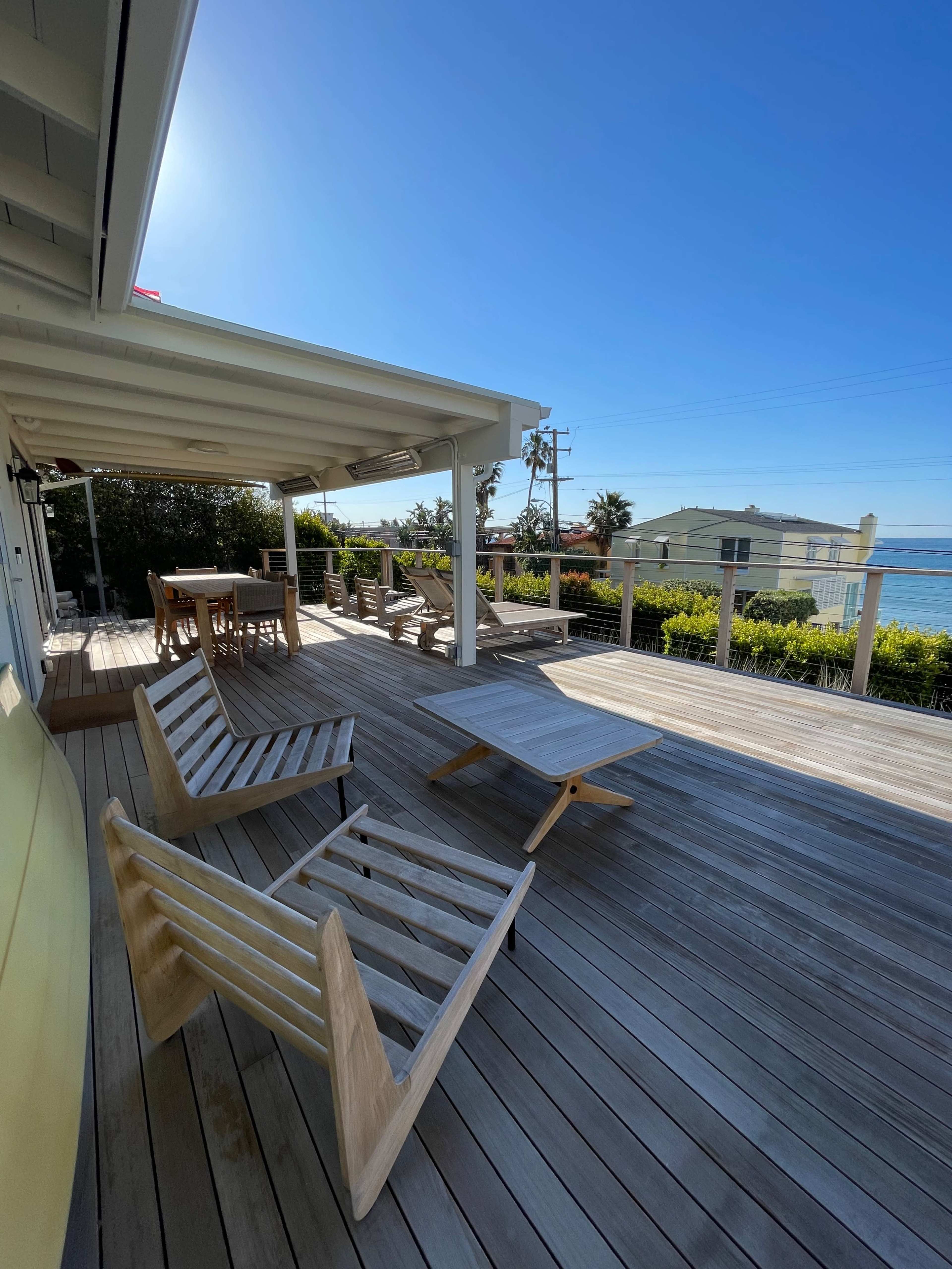 A wooden deck features lounge chairs and a dining area with a view of the ocean and nearby homes.