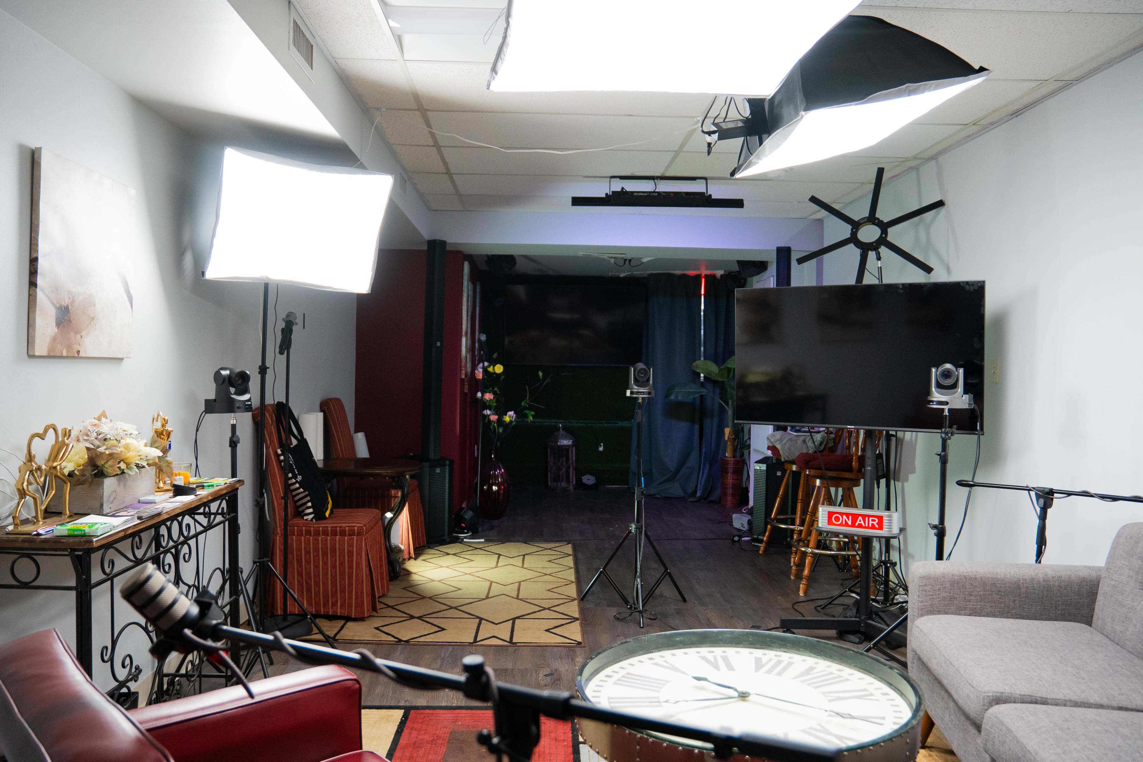 A recording studio setup featuring softbox lights, a large TV screen, a microphone, and a decorated seating area.
