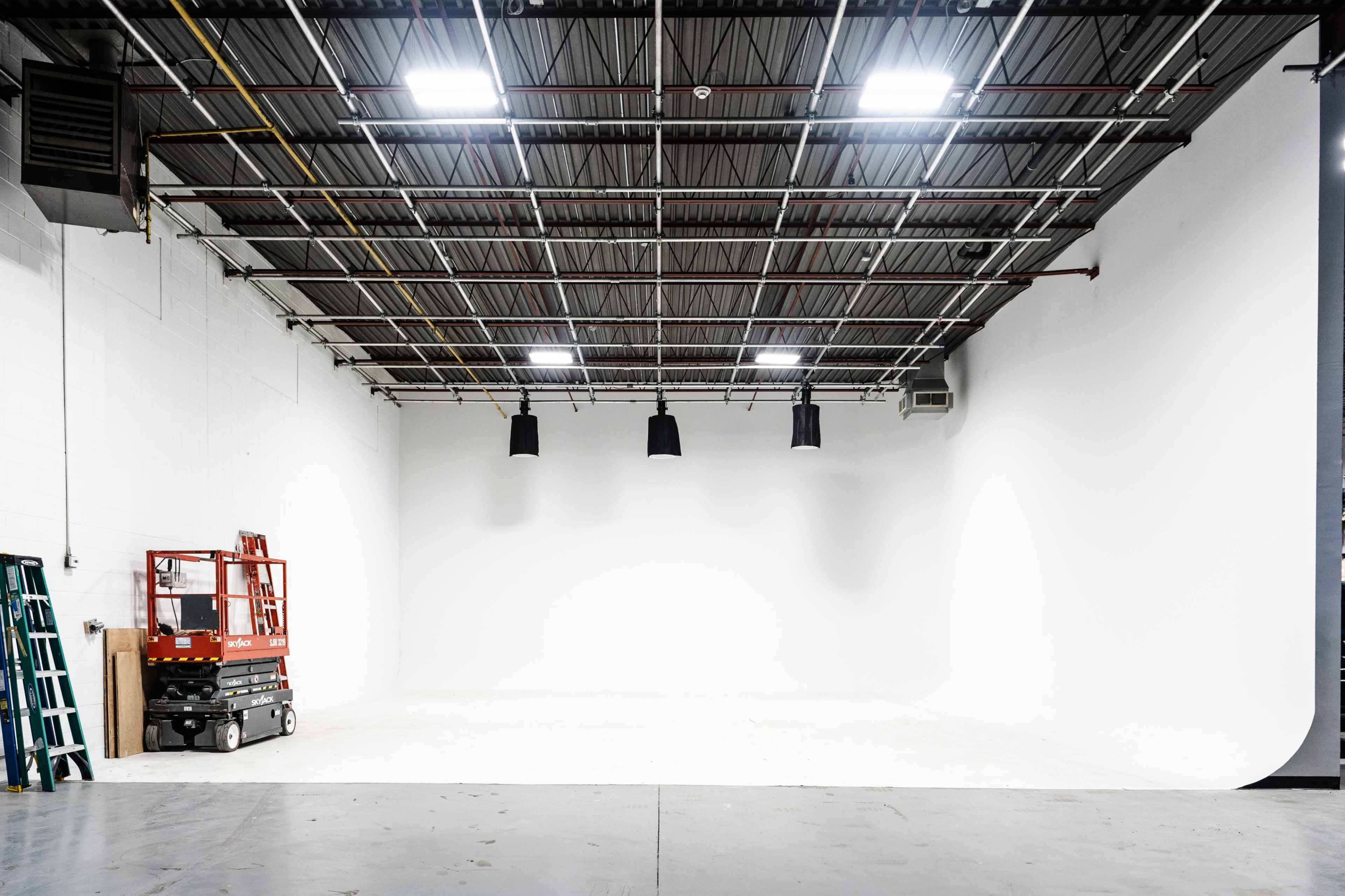 An empty studio space with a high ceiling, equipped with lighting fixtures and a scissor lift near a blank white backdrop.