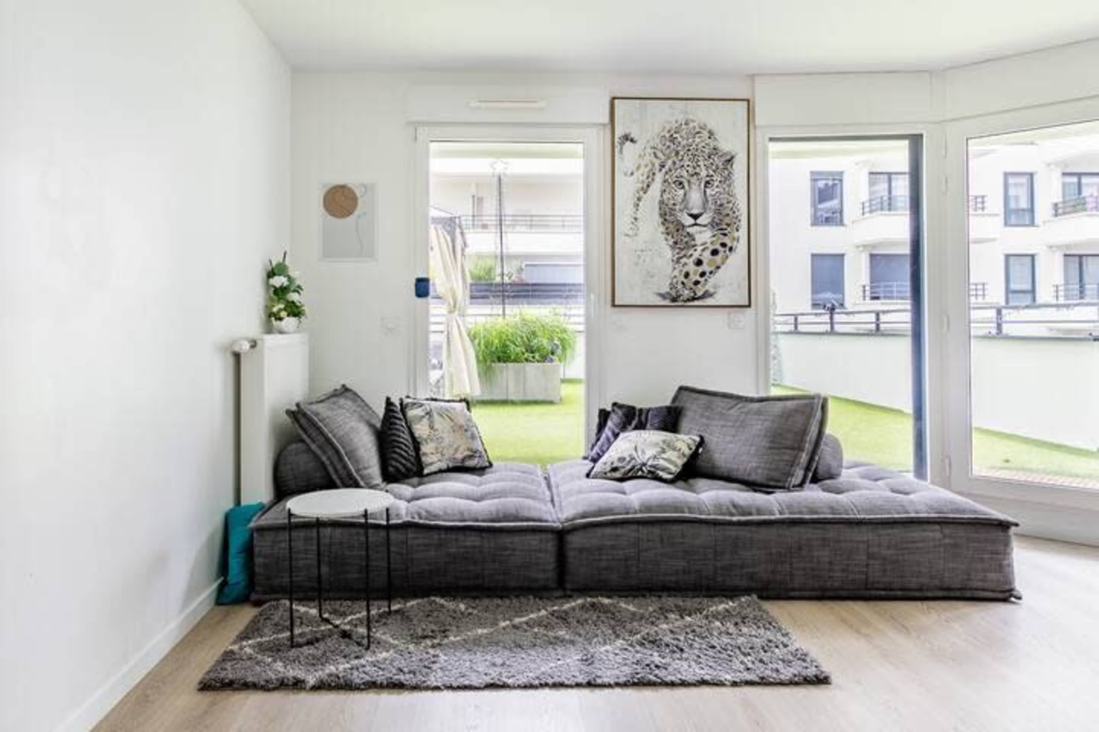 A modern living room features a large gray sofa with decorative pillows, a small side table, and an artwork of a leopard on the wall, with a view of a balcony garden through glass doors.