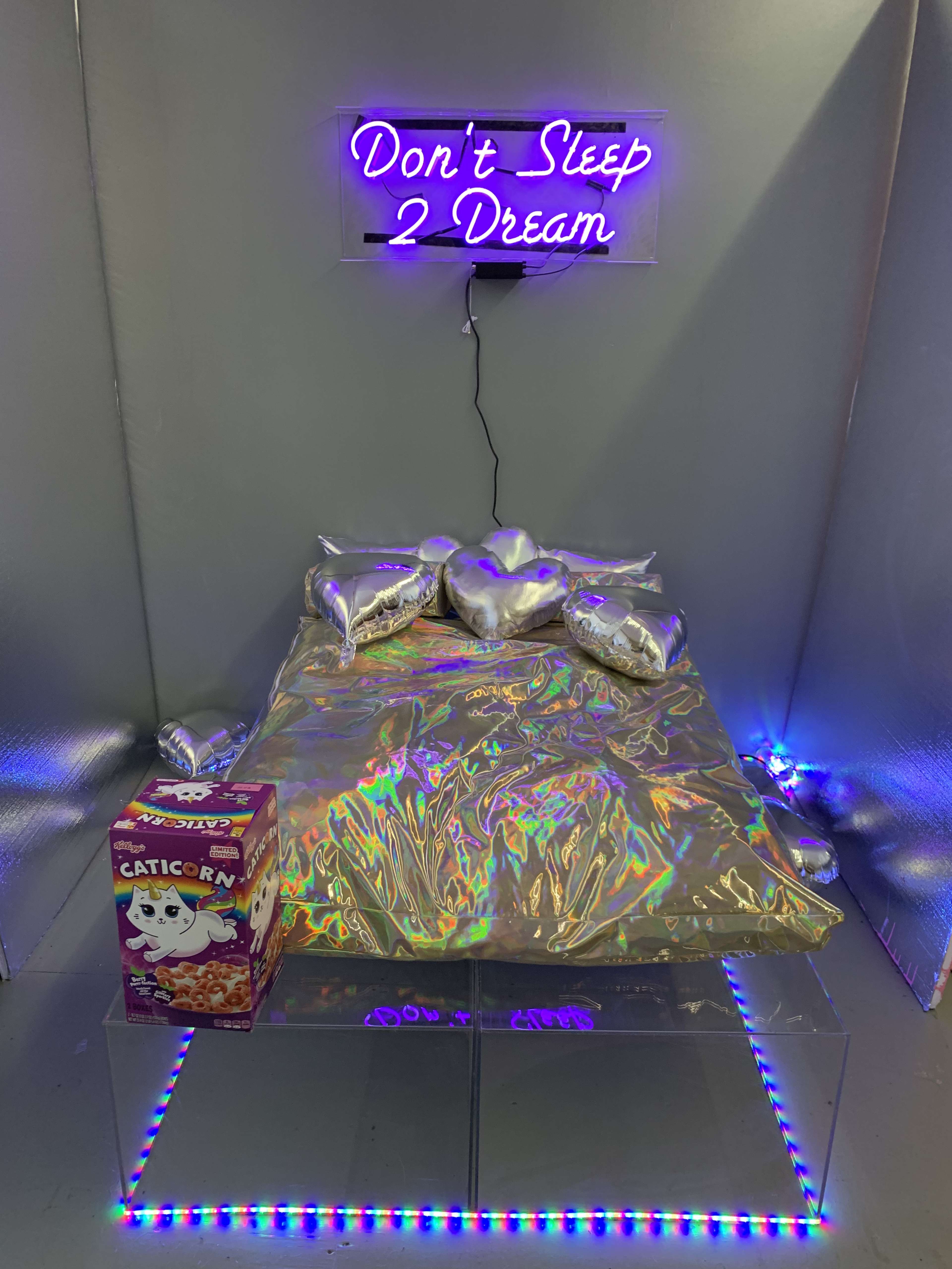 A bed with metallic silver pillows is placed under a neon sign that reads "Don't Sleep 2 Dream," beside a box of cereal.