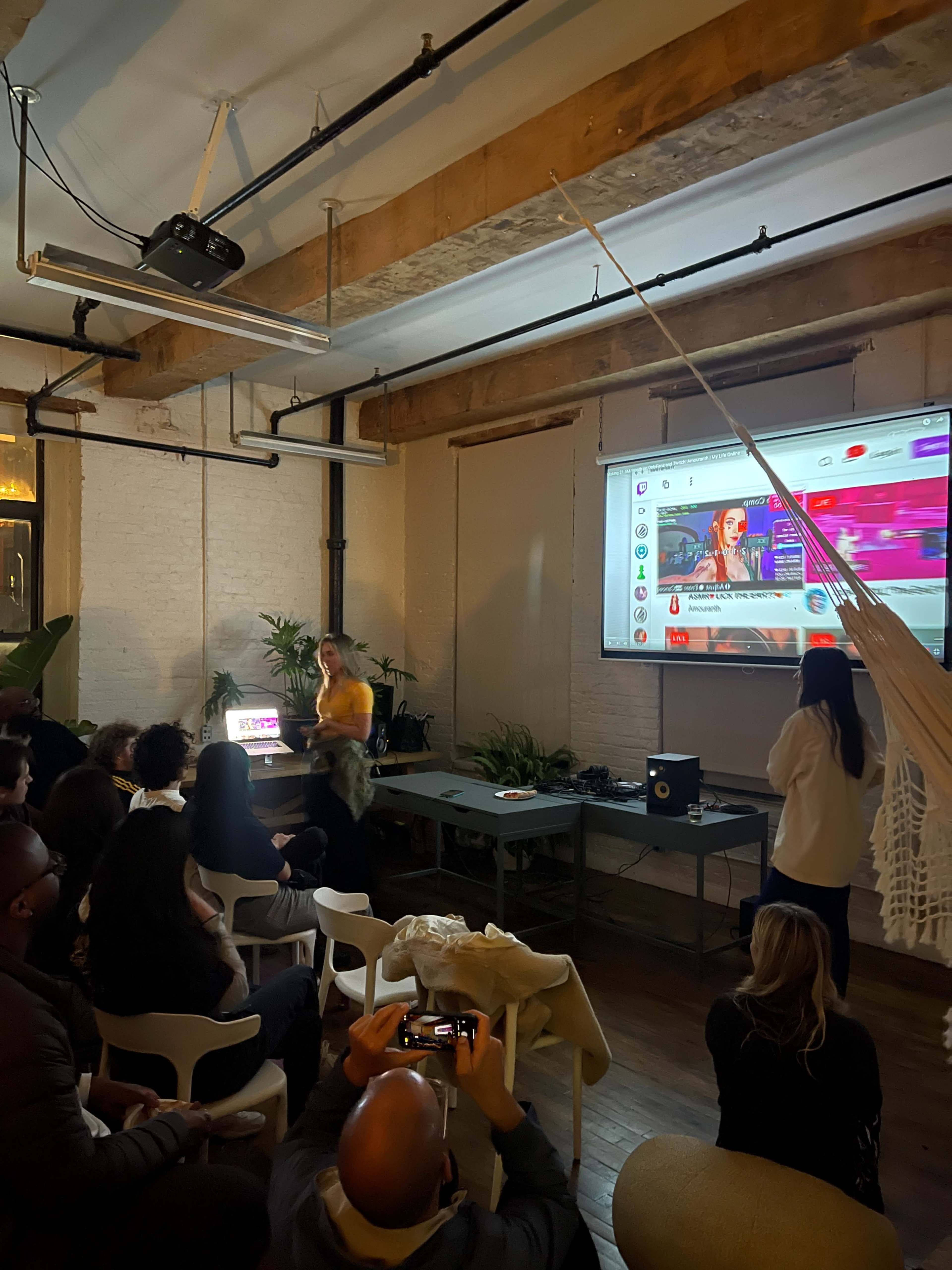 A speaker presents in front of an audience seated in a spacious room with a projector displaying digital content on a large screen.