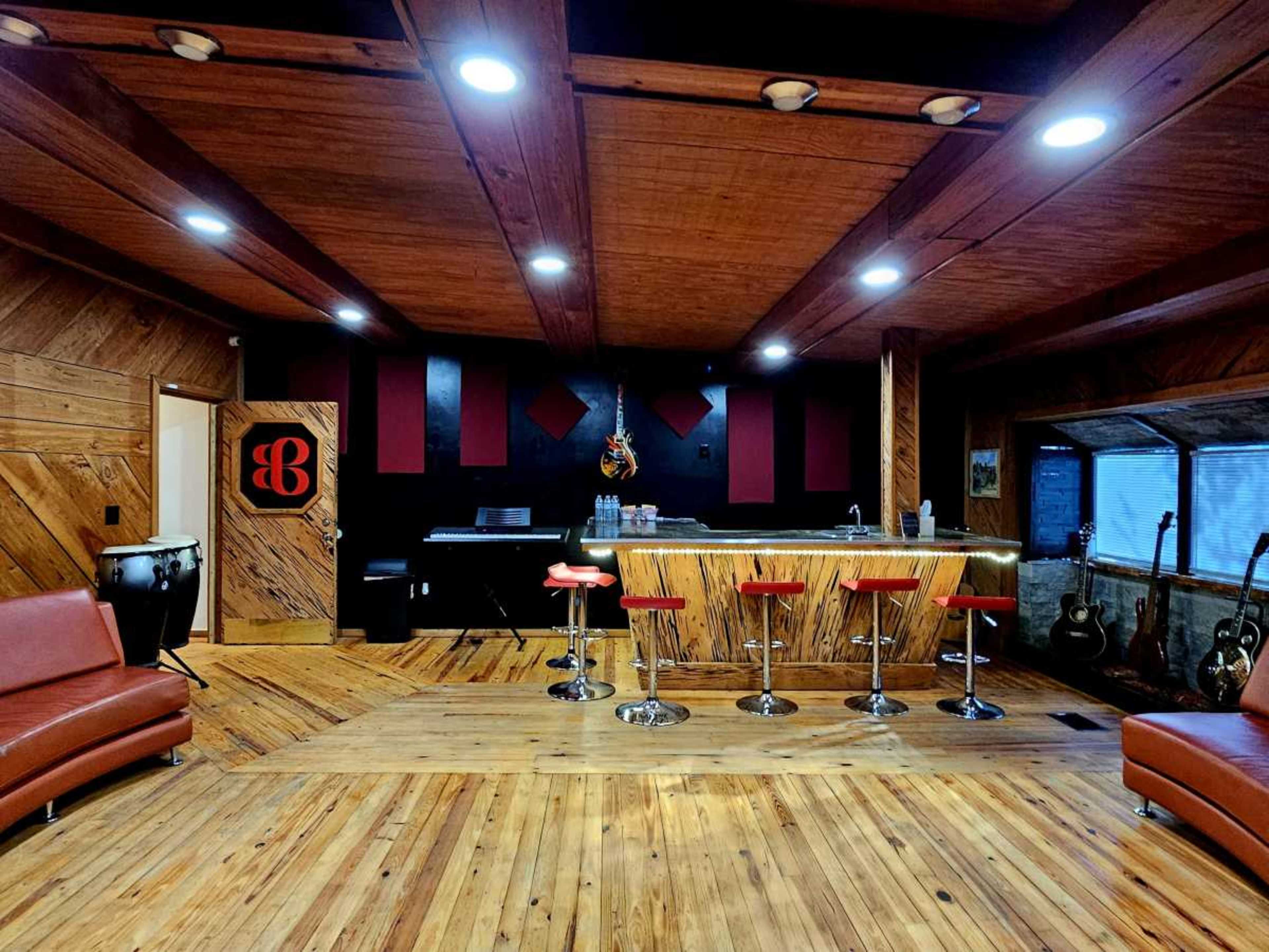 The image shows a wooden-paneled room featuring a bar area with red stools, a leather sofa, and musical instruments displayed in the corner.