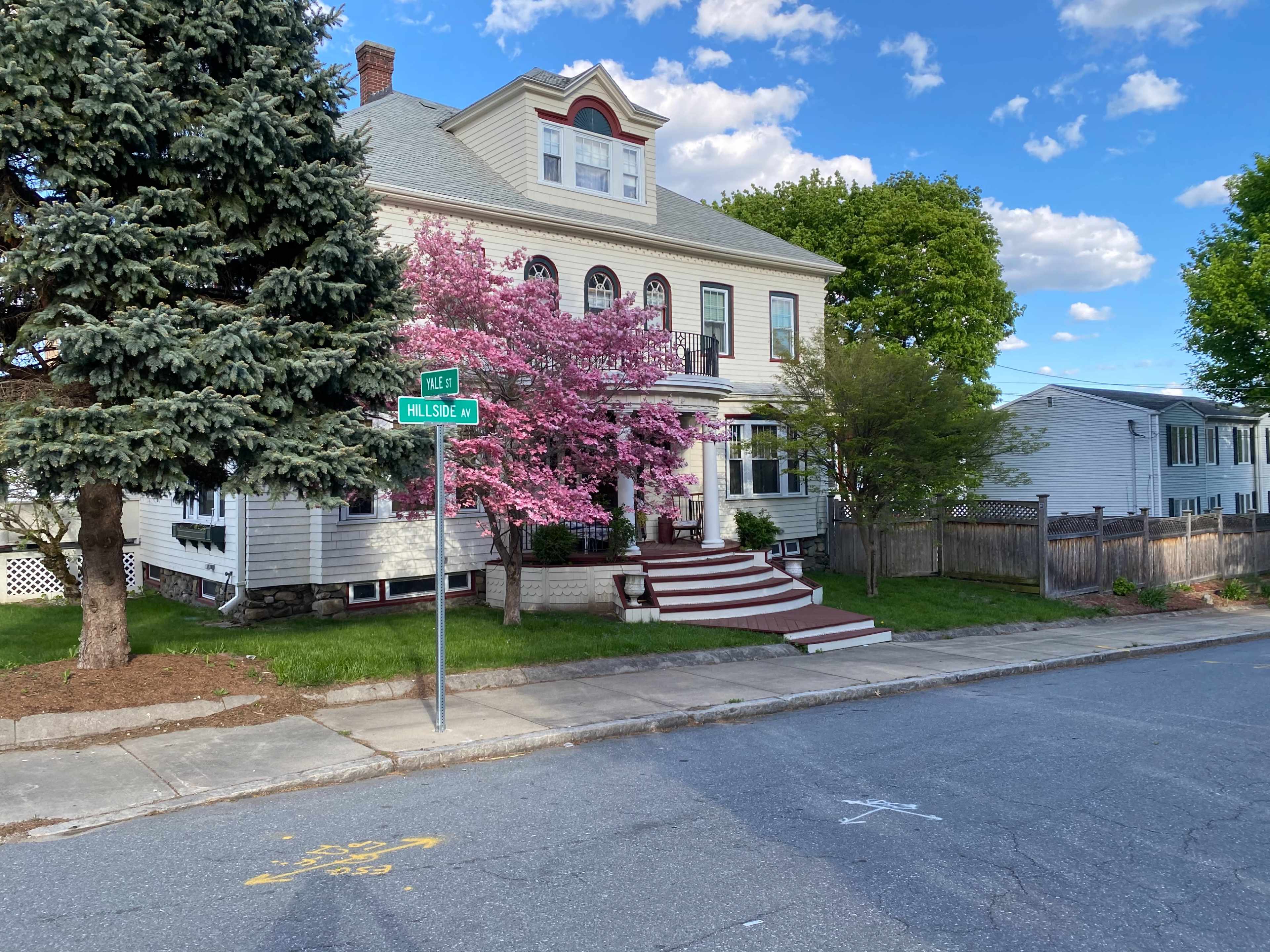 A large, two-story house with a gray roof and white siding is situated at the corner of a street, featuring a blooming pink tree in the front yard.