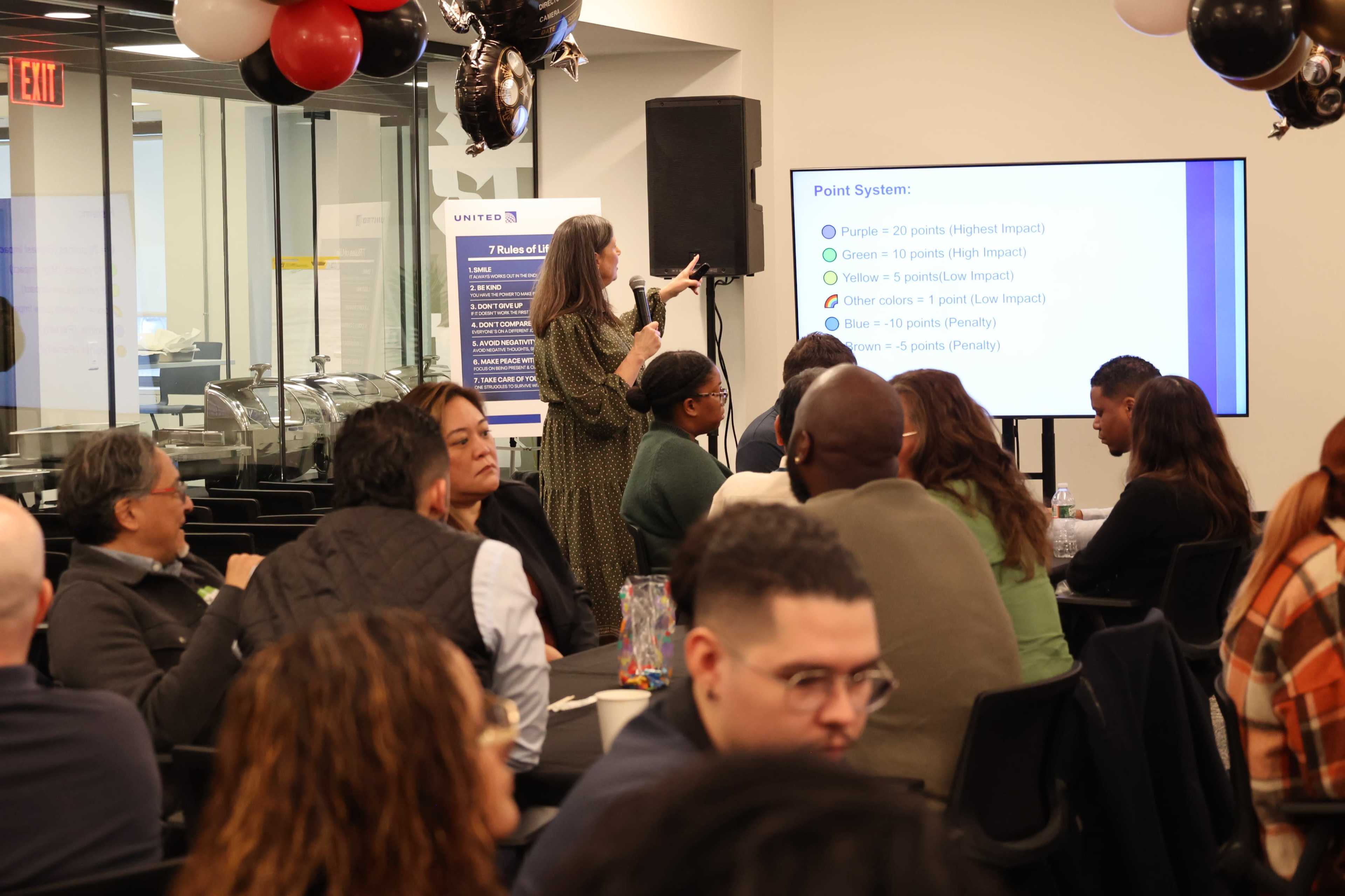 A woman presents information about a point system to an audience seated at tables in a conference room decorated with balloons.