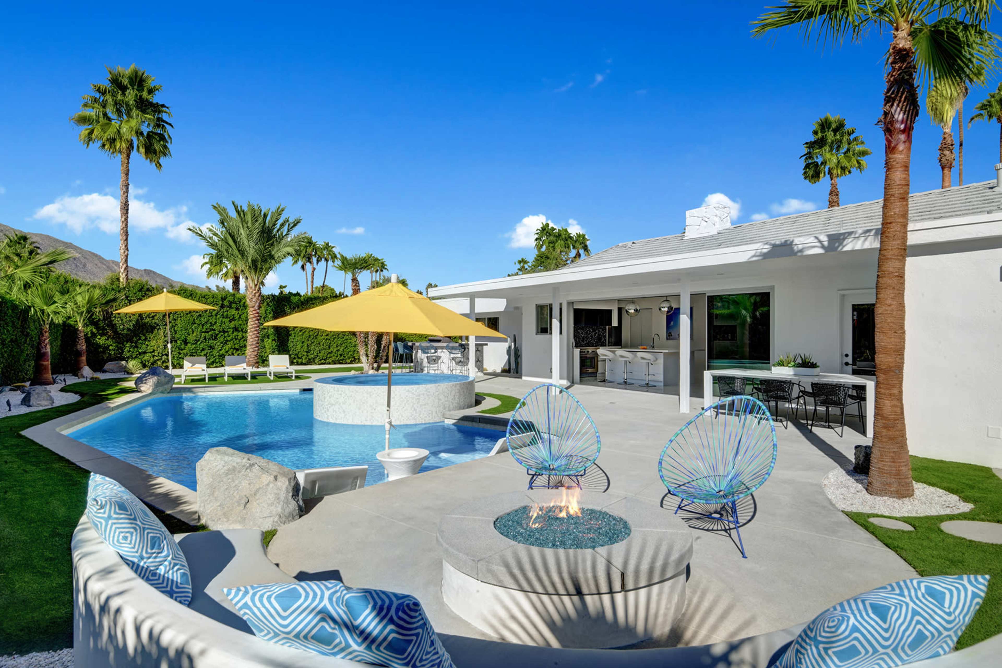 A modern backyard features a pool with lounge chairs, vibrant umbrellas, and a patio area with a fire pit surrounded by colorful chairs.