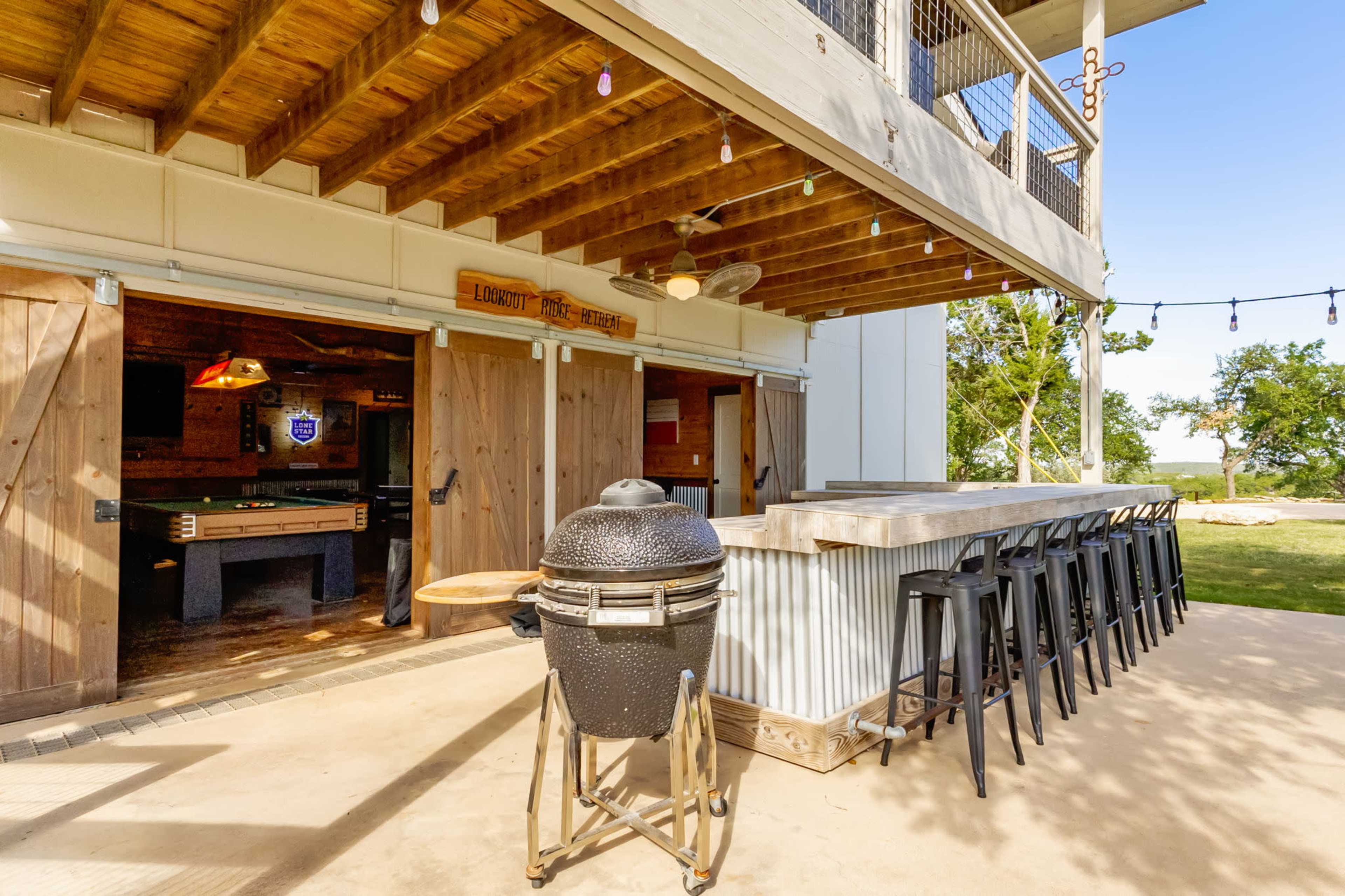 The image shows a covered outdoor area featuring a bar with high stools, a grill, and several wooden doors leading to an interior game space.