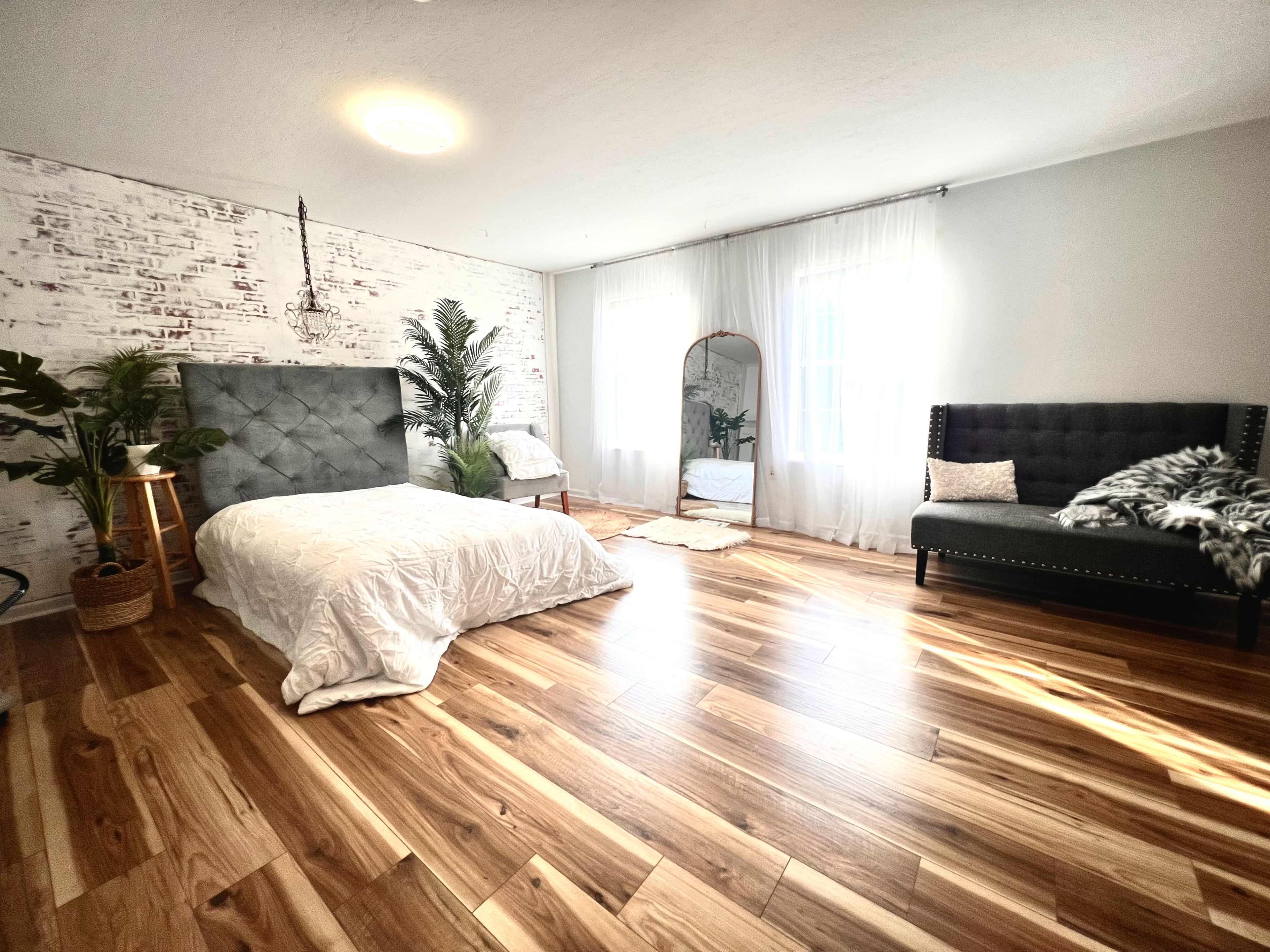 The room features a bed with a gray upholstered headboard, a large mirror, potted plants, and hardwood flooring, illuminated by natural light from two windows.