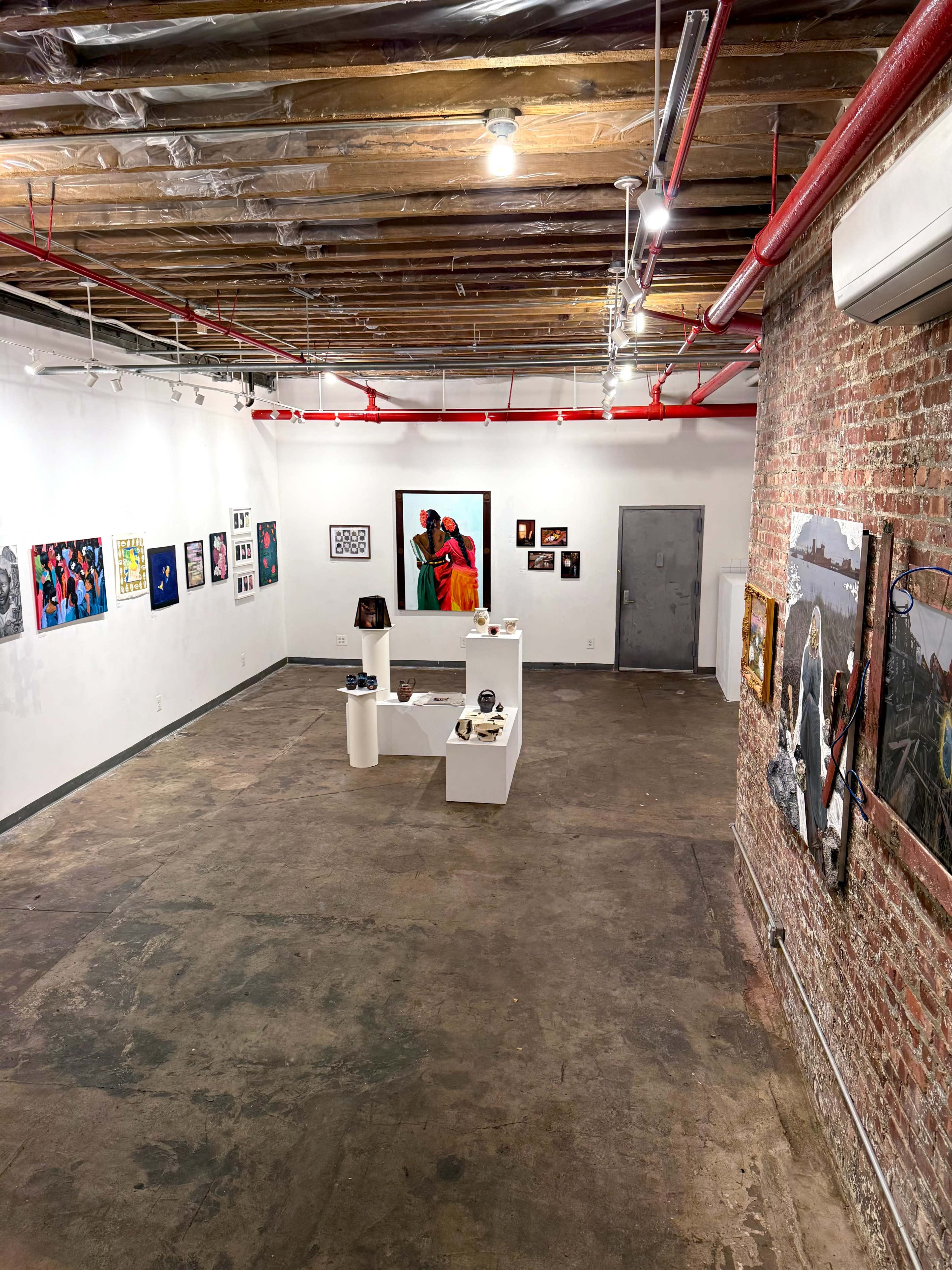 The image depicts an art gallery with several framed artworks hanging on the walls and a central display featuring sculptures or smaller pieces on white pedestals.