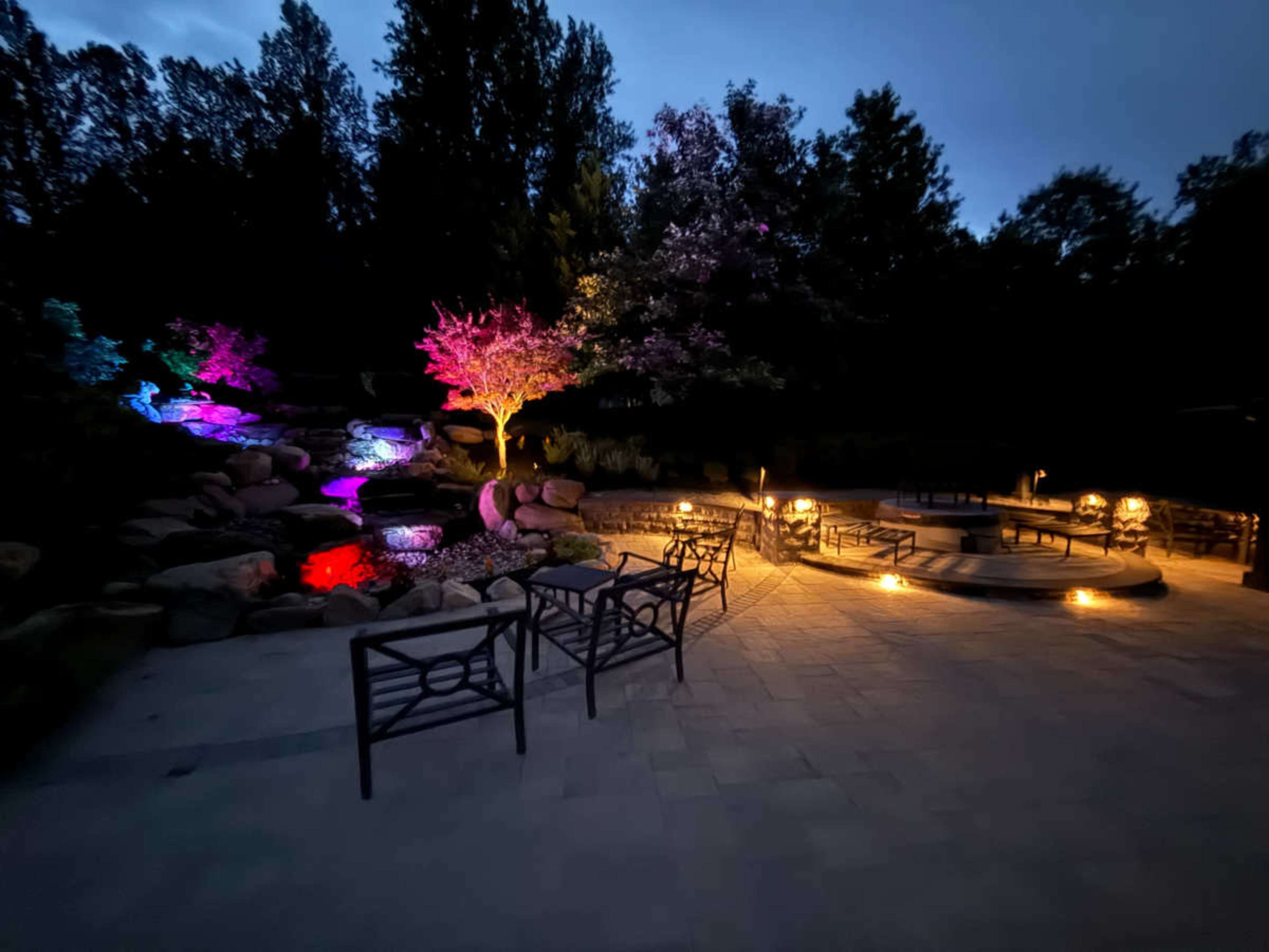 The image shows a landscaped outdoor area at dusk, featuring colorful lights highlighting trees and a pond, with seating arranged around a circular fire pit.