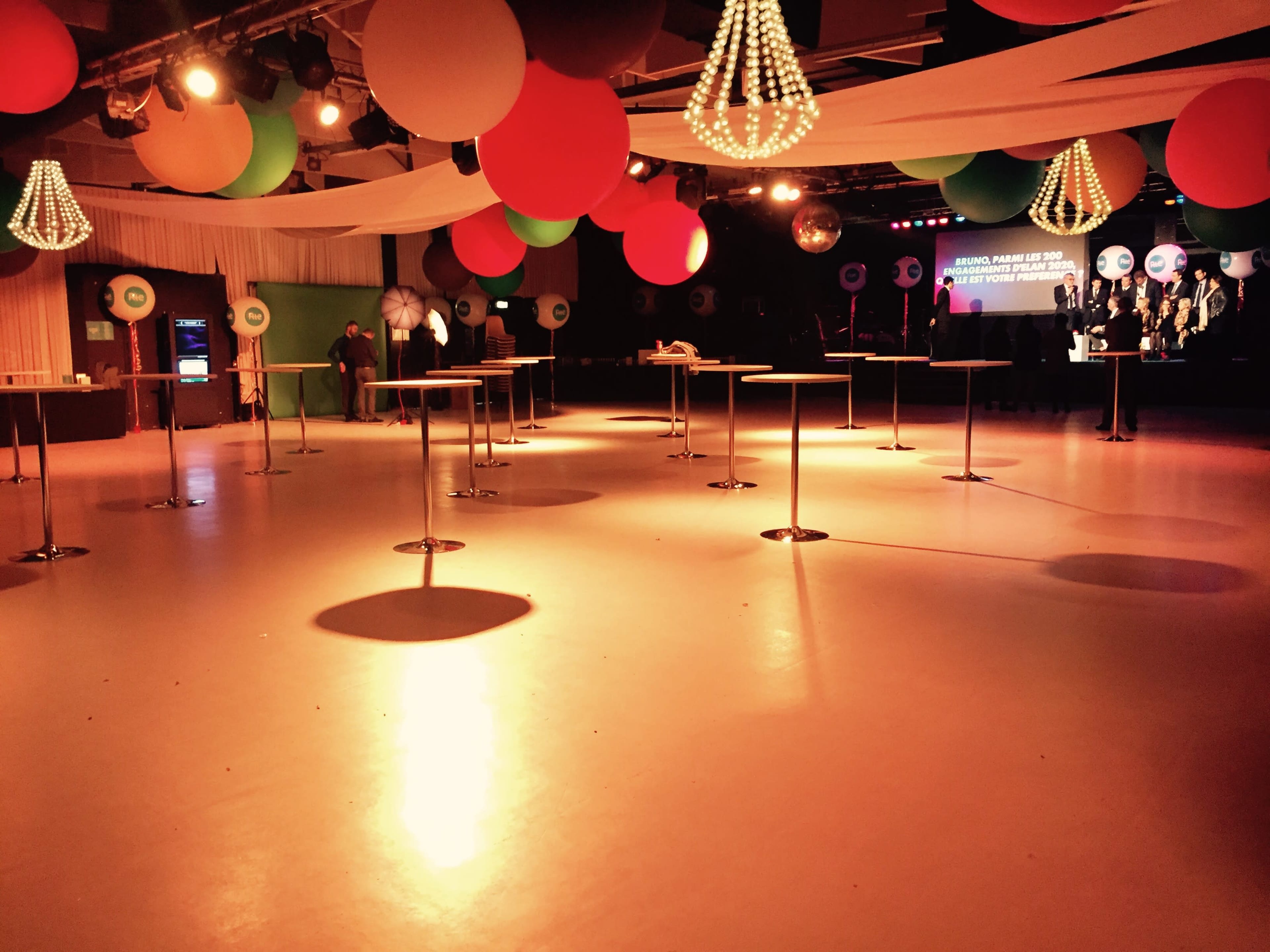 An empty event space with round tables, colorful hanging decorations, and a stage at the back.