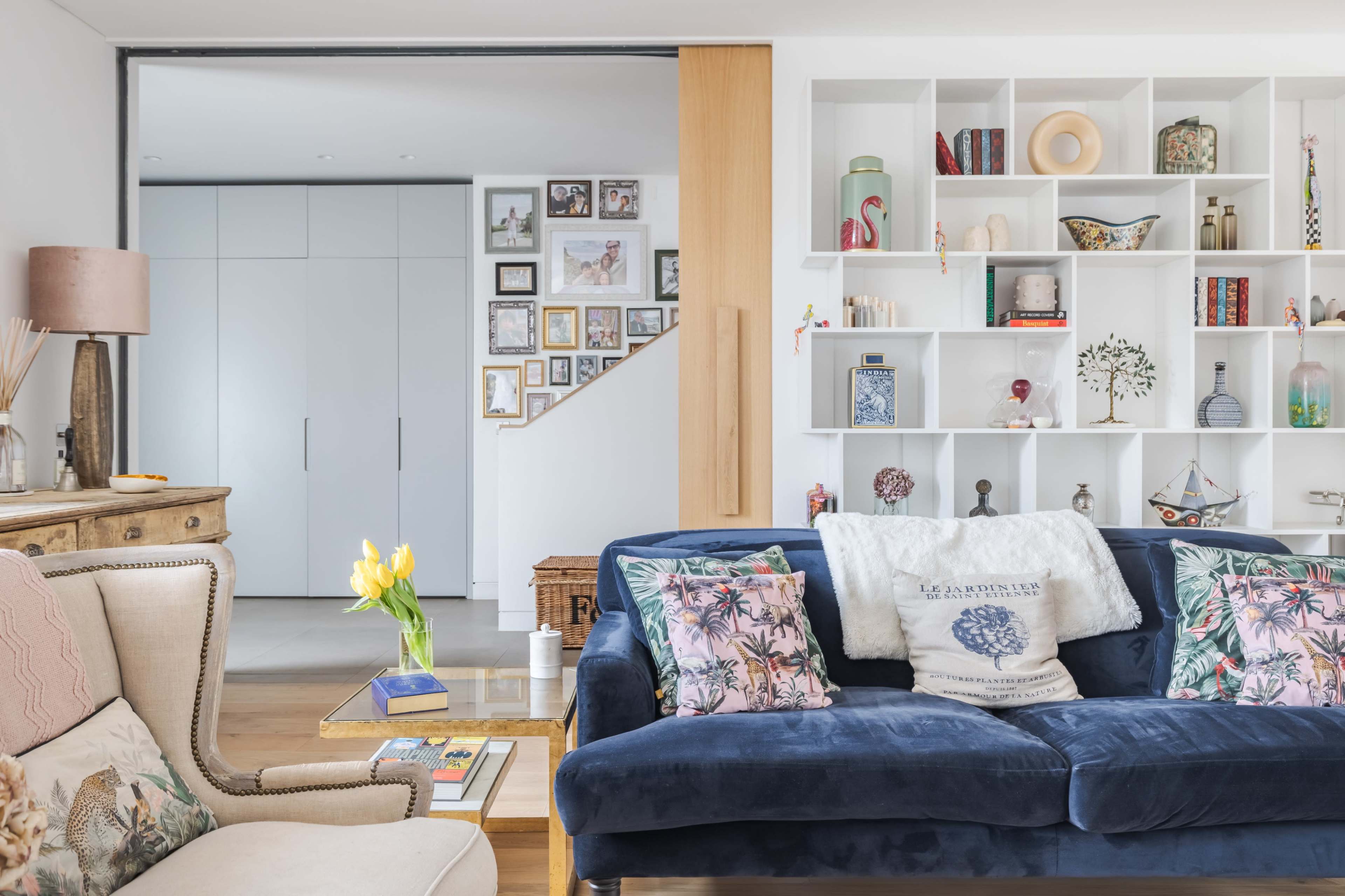 The image shows a modern living room with a blue sofa, decorative pillows, and a wall unit displaying various decorative items and framed photographs.