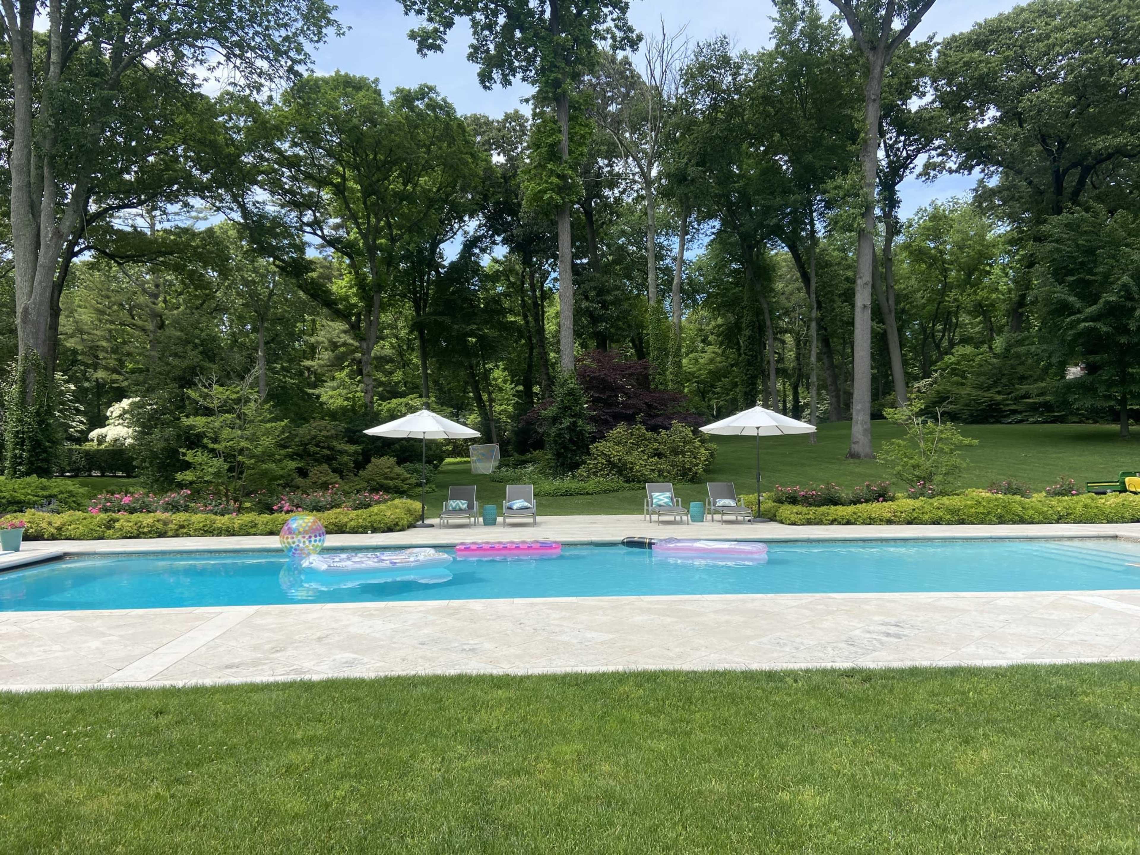 The image shows a swimming pool surrounded by green grass and trees, with floating pool floats and lounge chairs under umbrellas on the deck.