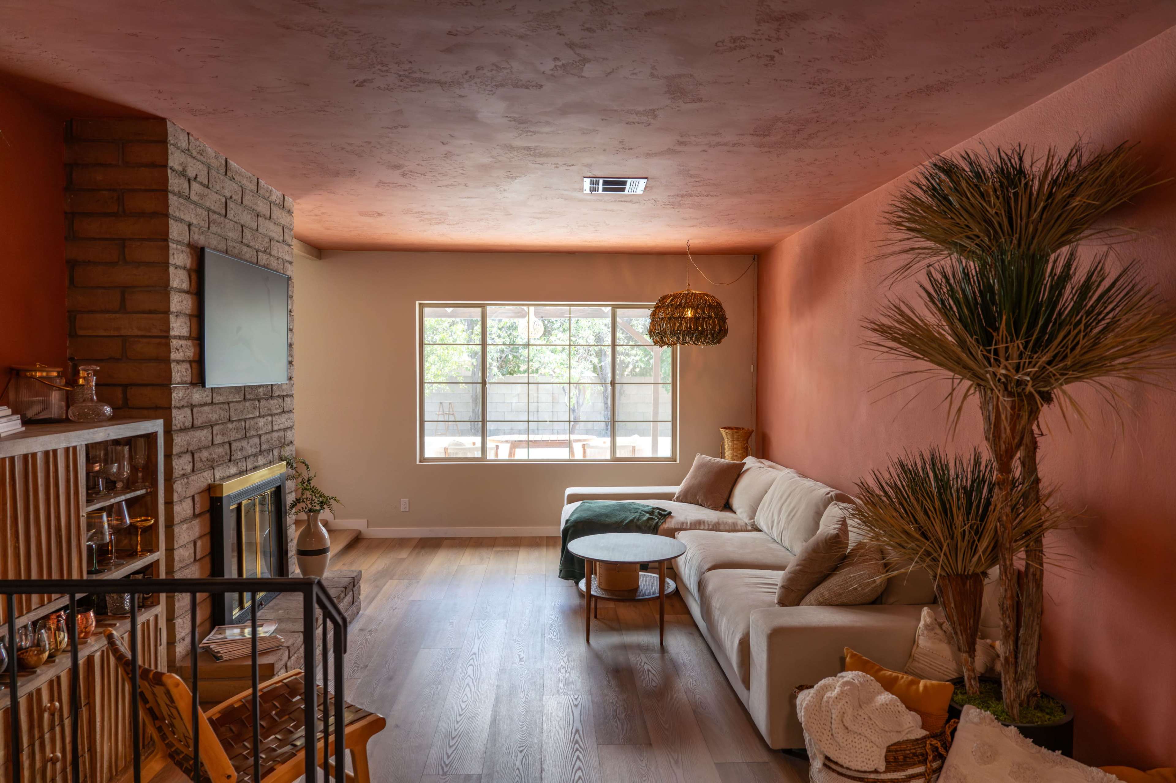 The image shows a living room featuring a beige sofa, a wooden coffee table, a stone fireplace, and decorative plants, with a large window allowing natural light to enter.