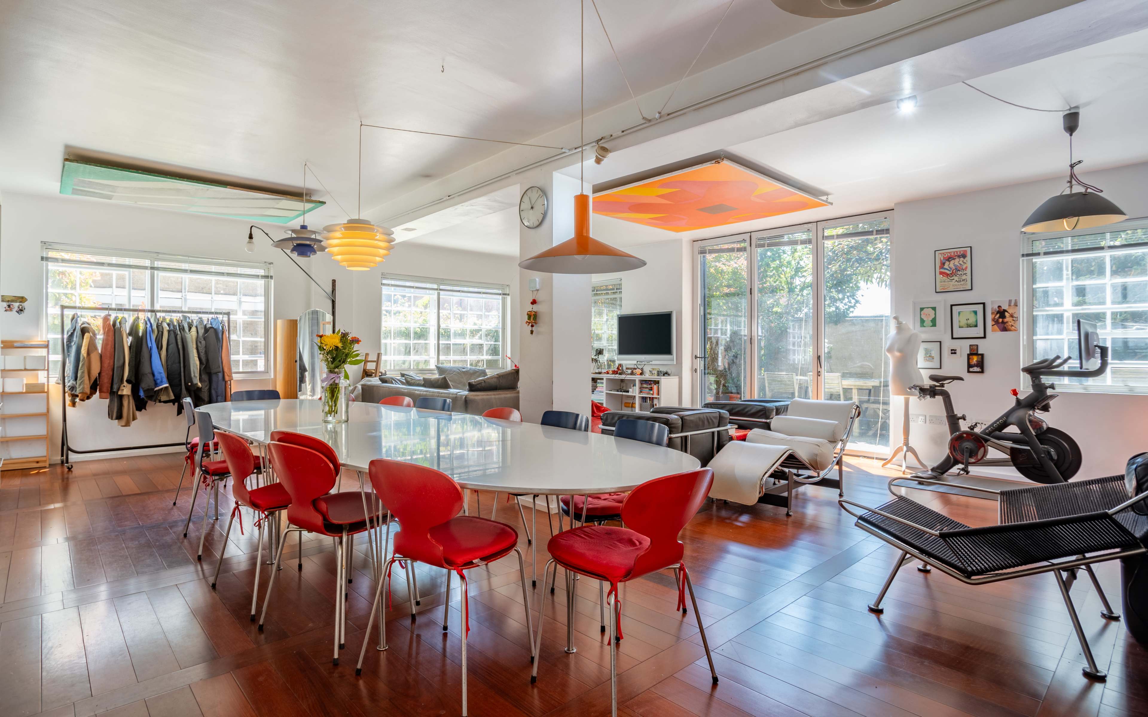 The image shows a spacious modern living area with a large dining table surrounded by red chairs, a lounge area with sofas, and various decorative items and plants.
