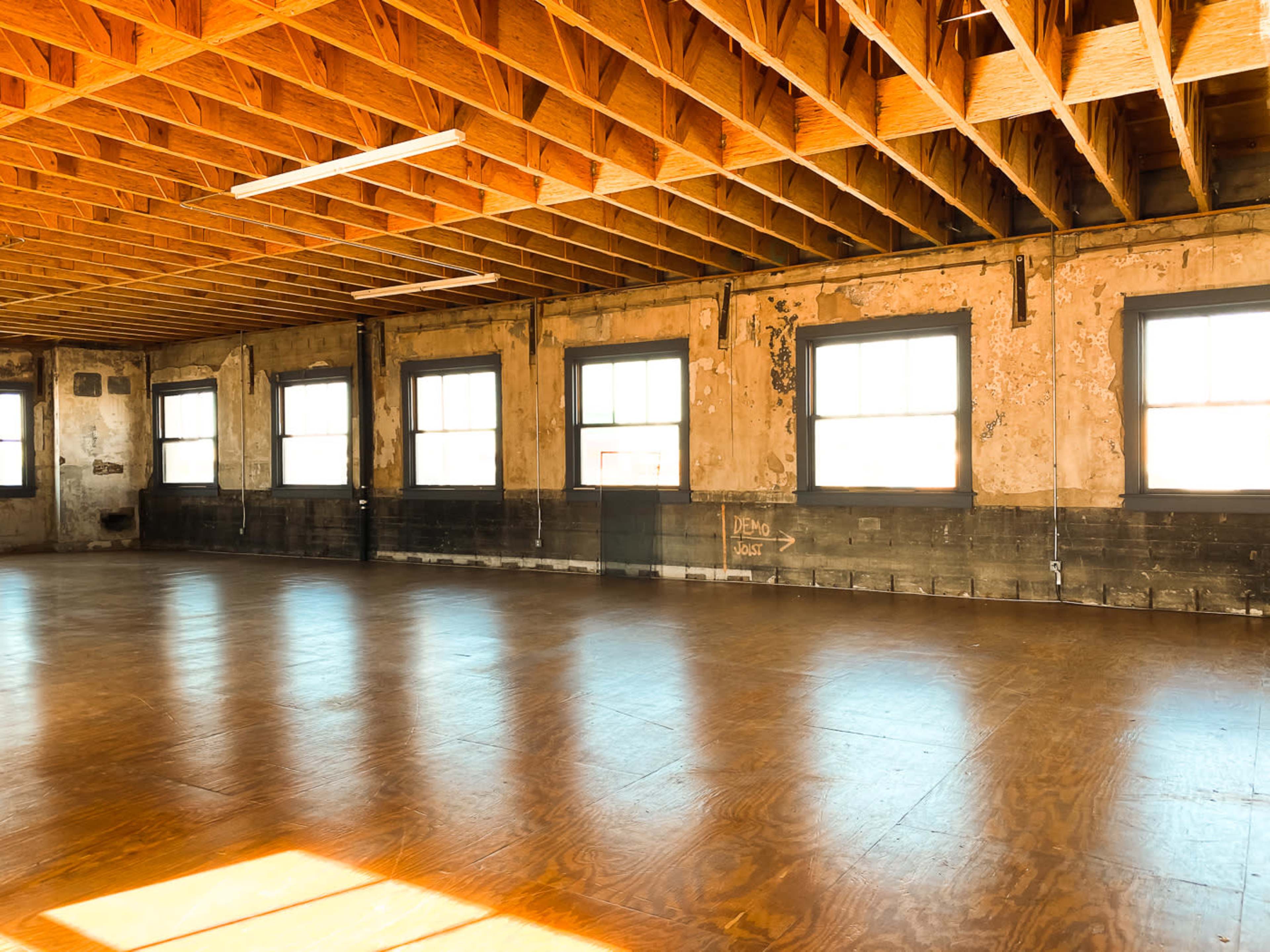 The image shows a spacious, empty room with wooden floors and large windows, characterized by exposed beams and a weathered interior.