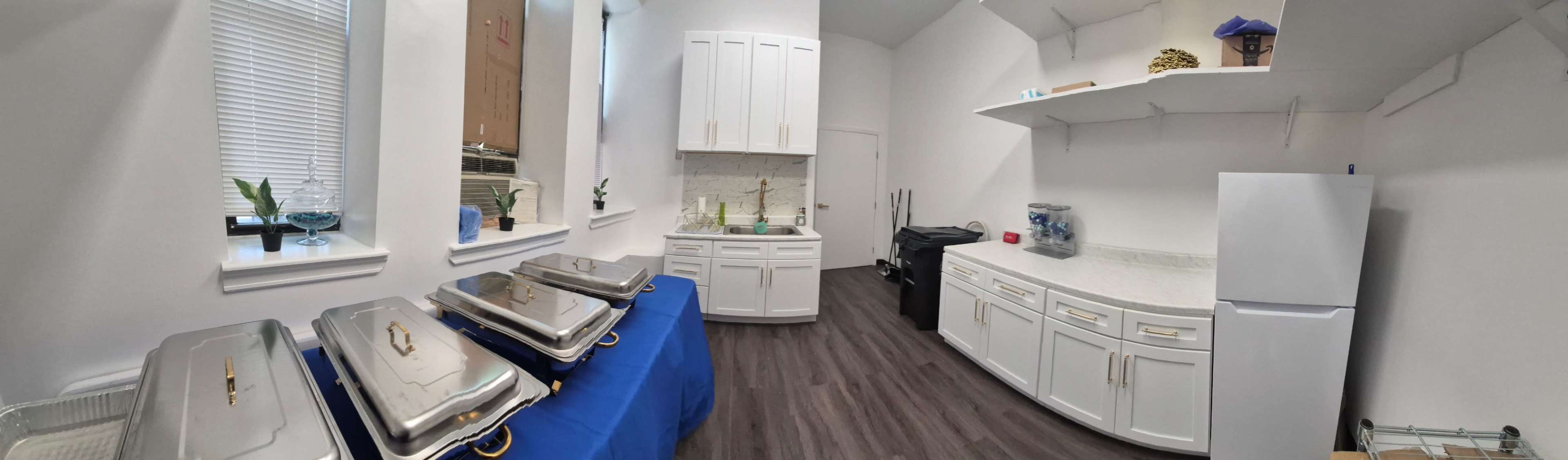 The image shows a bright kitchen featuring white cabinetry, a refrigerator, and multiple chafing dishes on a blue table.