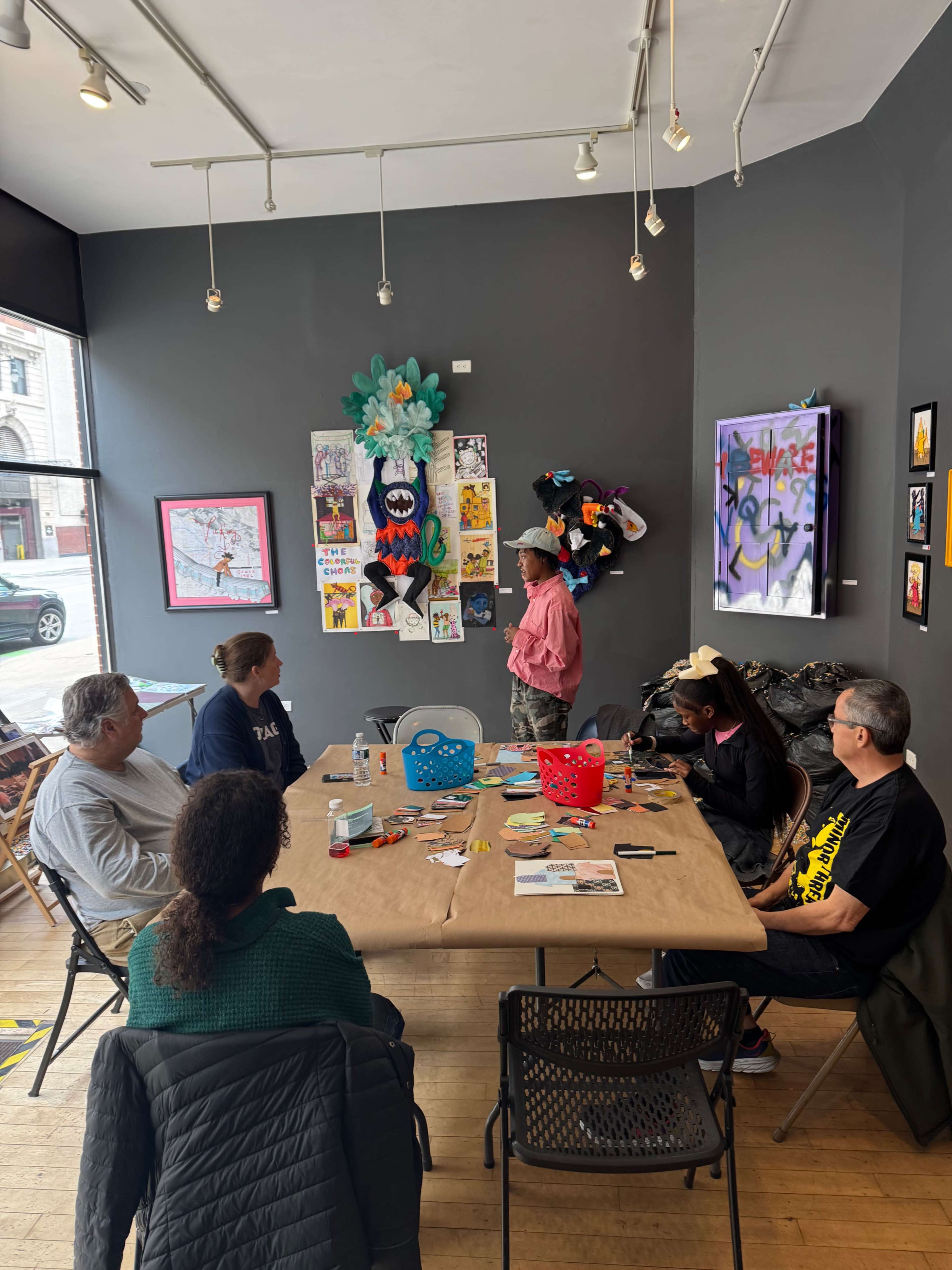 A group of six people sits around a table in an art gallery, with colorful artwork displayed on the walls and someone standing in front of them speaking.