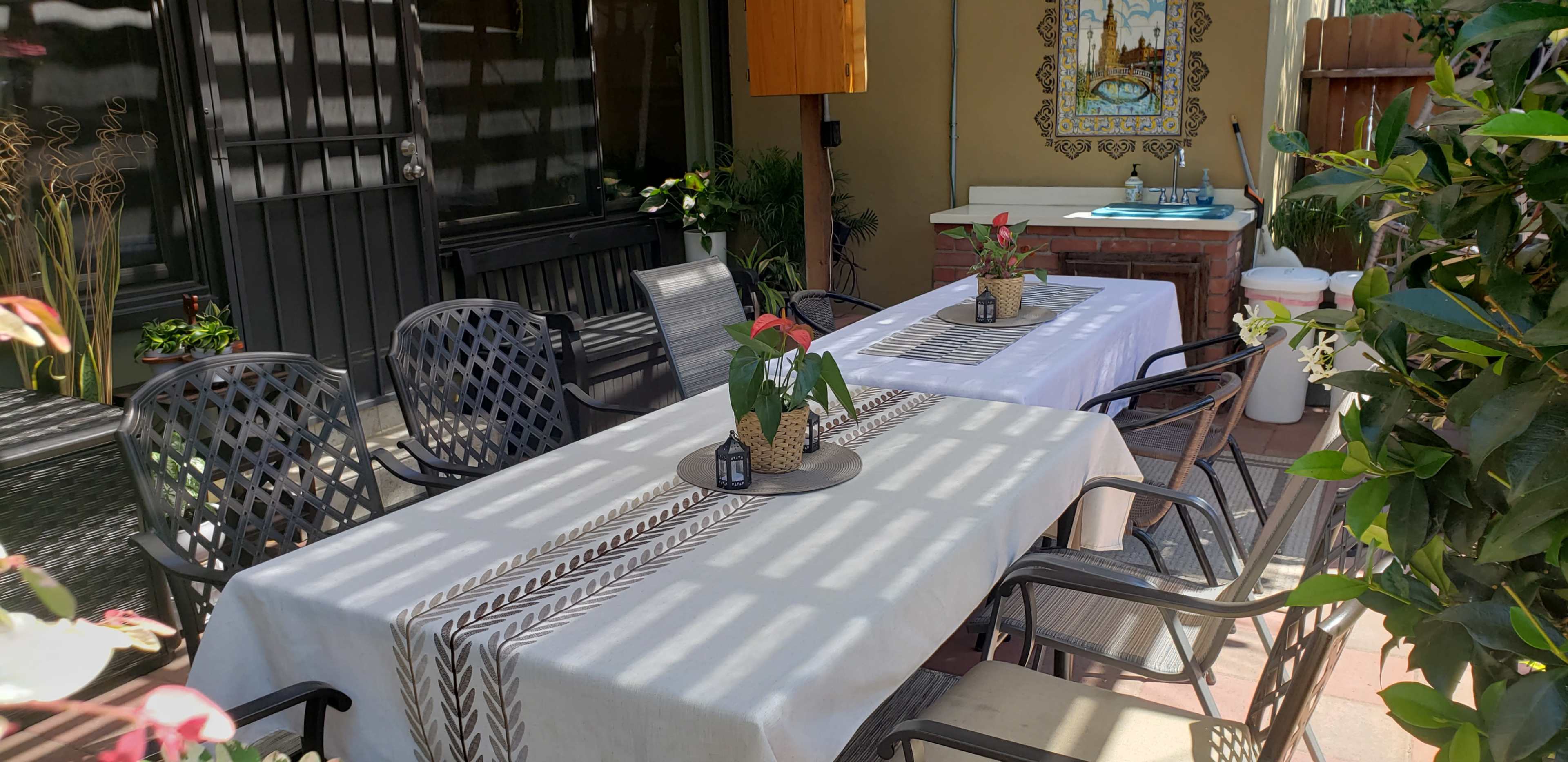 The scene features a dining area with two long tables set with beige tablecloths, surrounded by black metal chairs and adorned with potted plants.