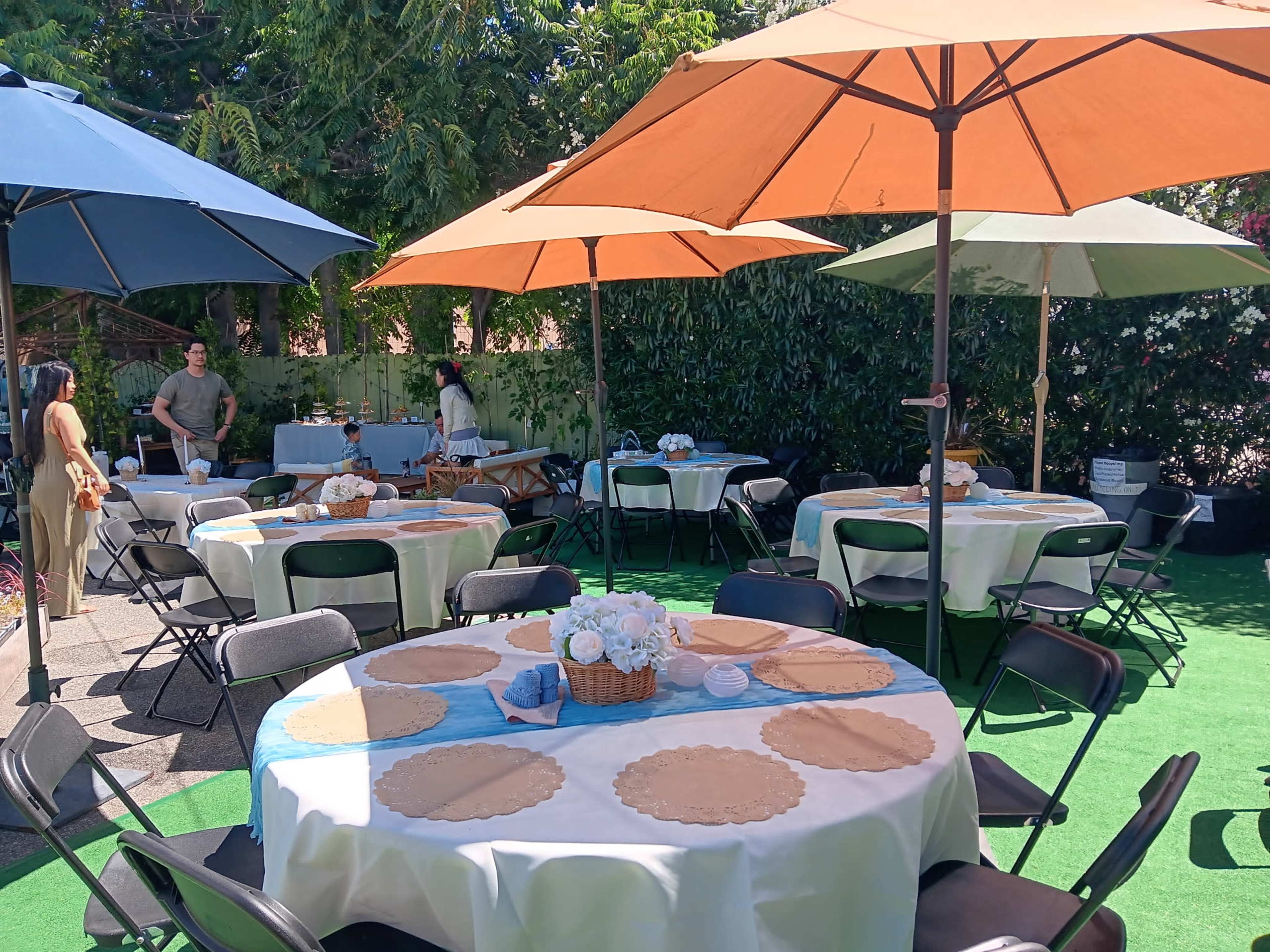 The image shows a garden party setup with multiple round tables covered in tablecloths, each decorated with centerpieces and shaded by large umbrellas.
