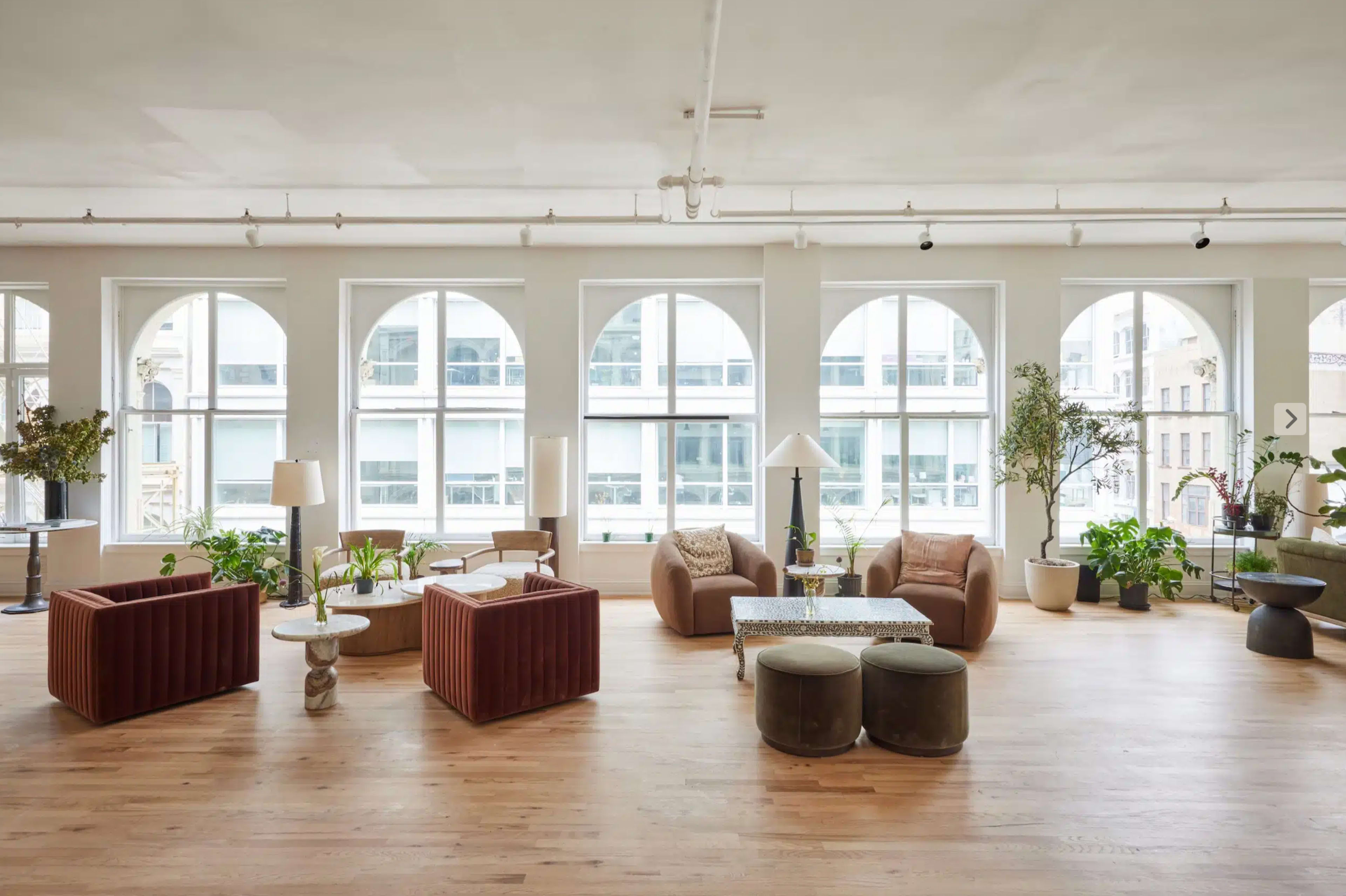 The image shows a spacious, well-lit room with large arched windows, featuring a mix of modern seating arrangements, plants, and decorative tables on a wooden floor.