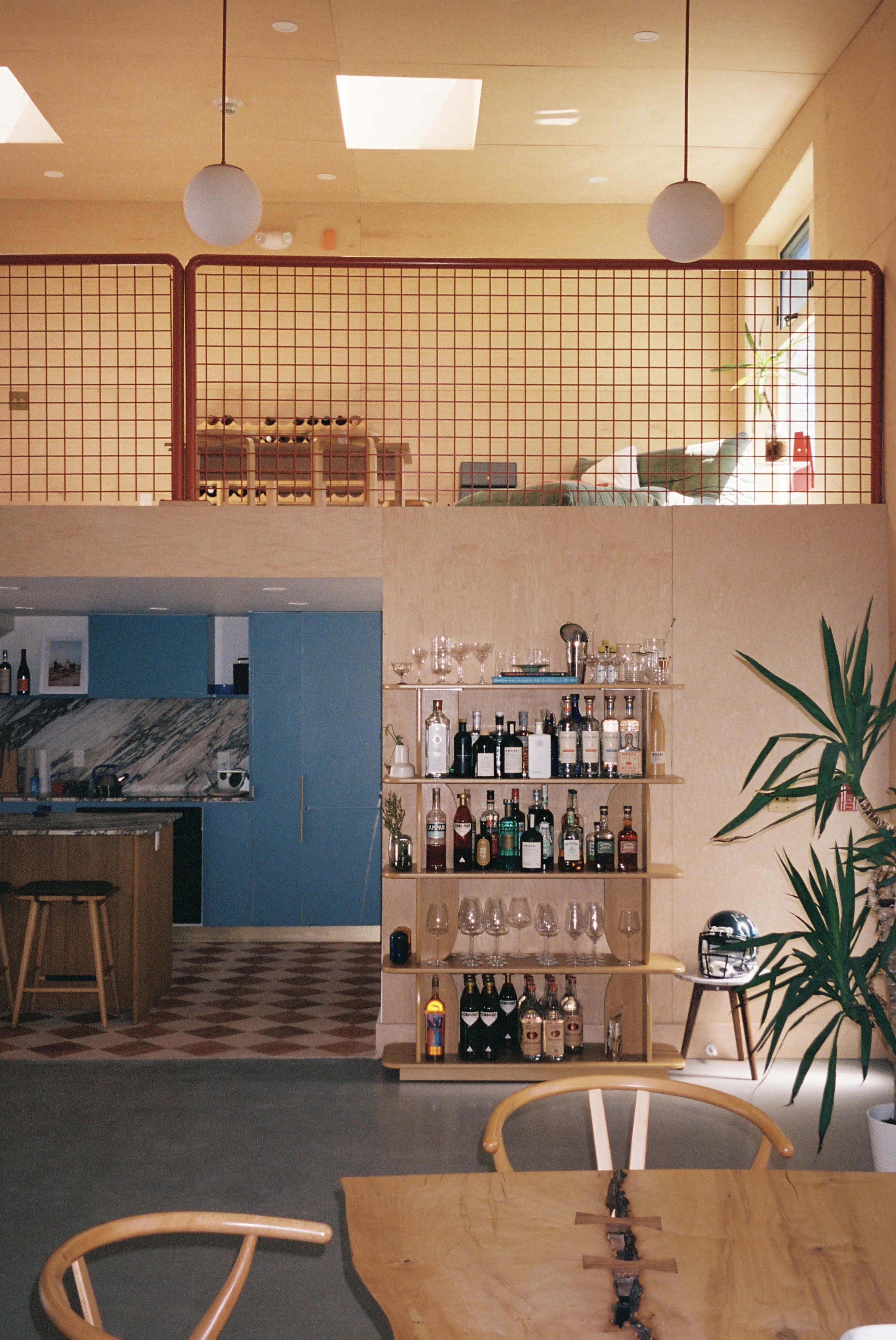 The image shows a modern interior of a home with a two-level space, featuring a wooden bar shelf filled with various bottles and glasses, and an open kitchen area in the background.