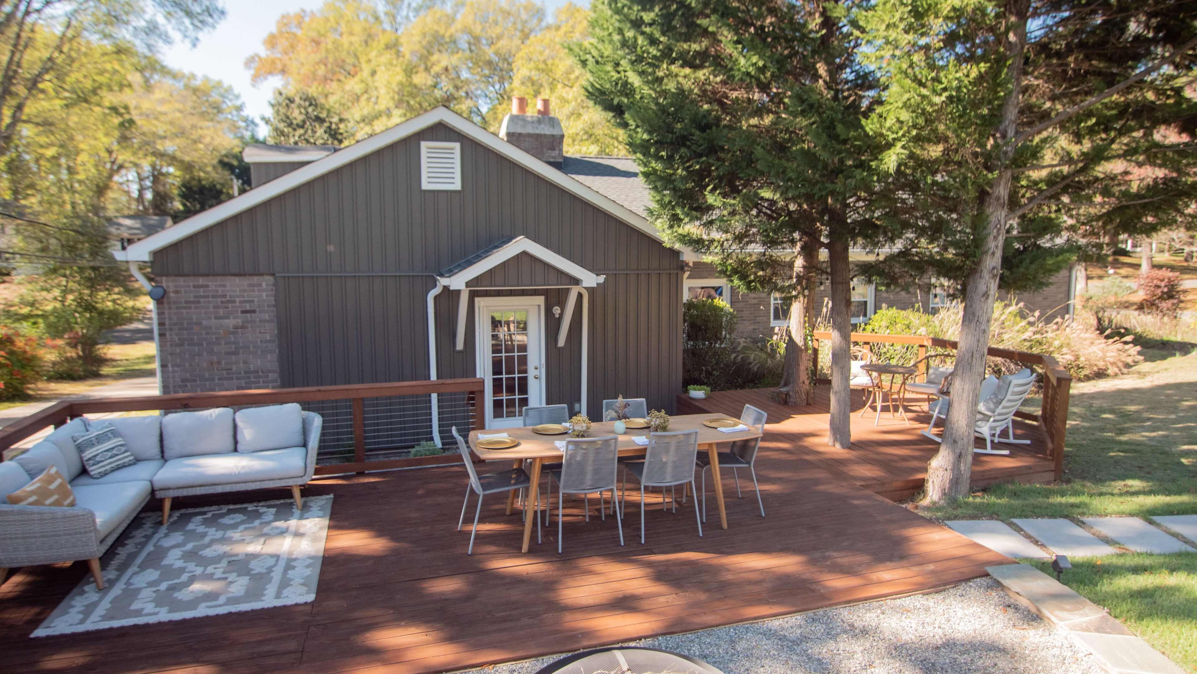 A wooden deck with a dining table and seating area is adjacent to a gray house surrounded by greenery.