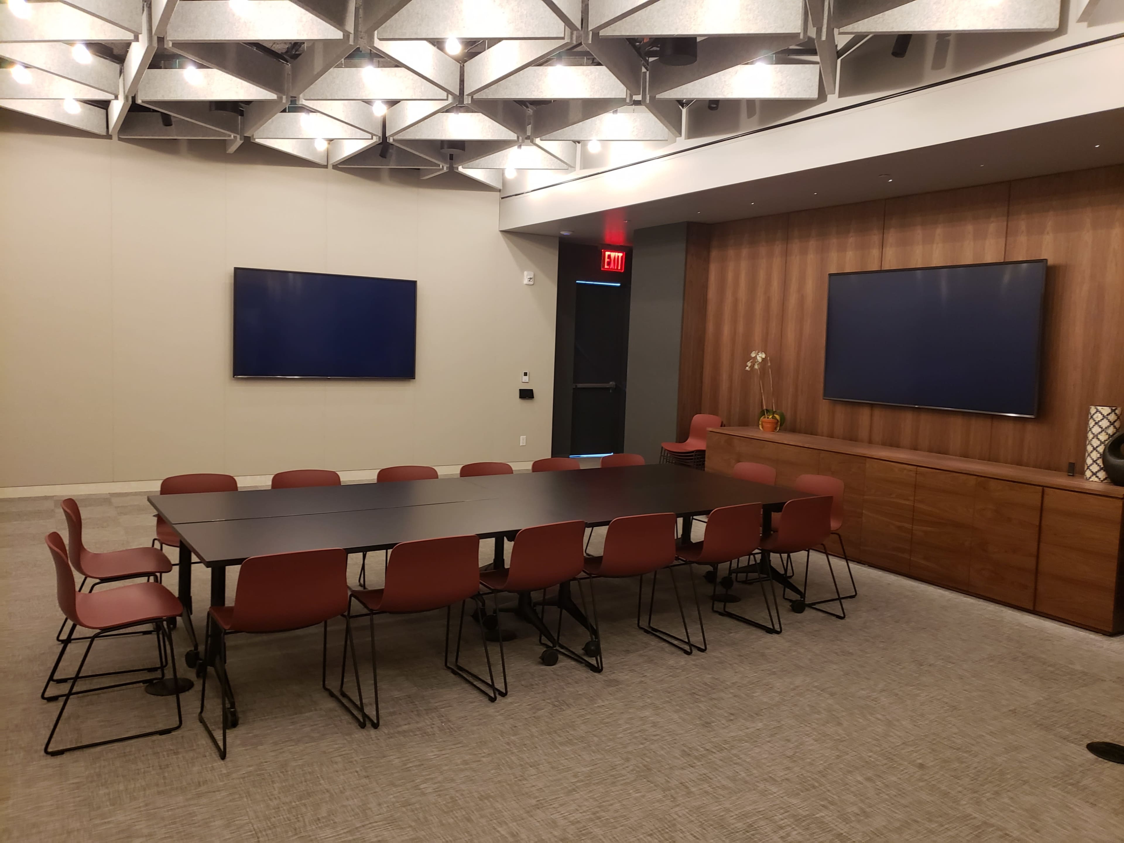 A modern conference room with a rectangular table surrounded by twelve chairs, two large screens mounted on the walls, and wooden accents.