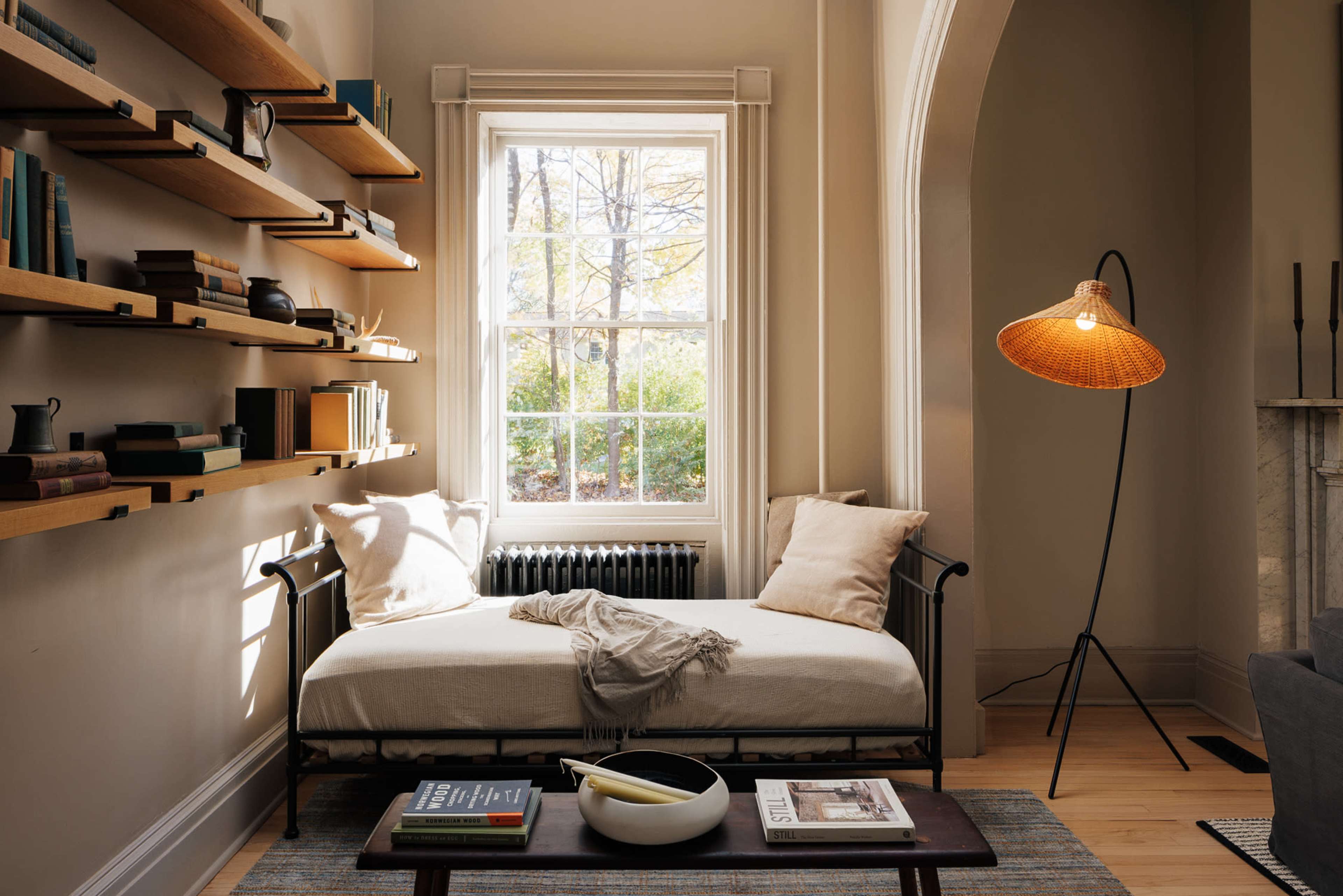 A cozy interior features a daybed beside a window, with wooden shelves displaying books and decorative items, and a floor lamp illuminating the space.